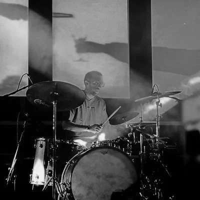 Un musicista che suona la batteria durante una performance, con luci e ombre evidenti sul palco.