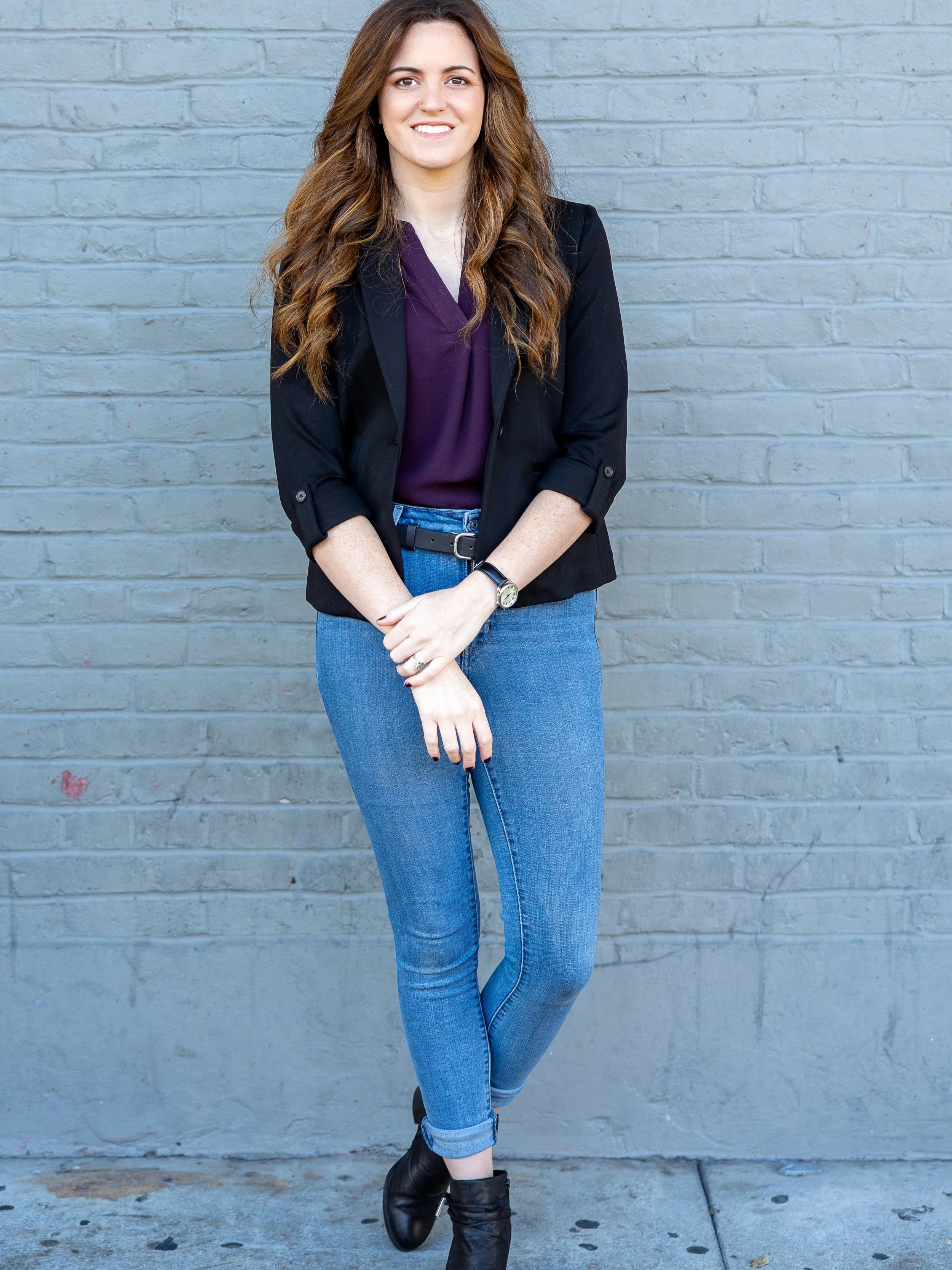 A young woman with long wavy brown hair, wearing a black blazer, purple top, blue jeans, and black ankle boots, standing against a gray brick wall, smiling and looking at the camera.