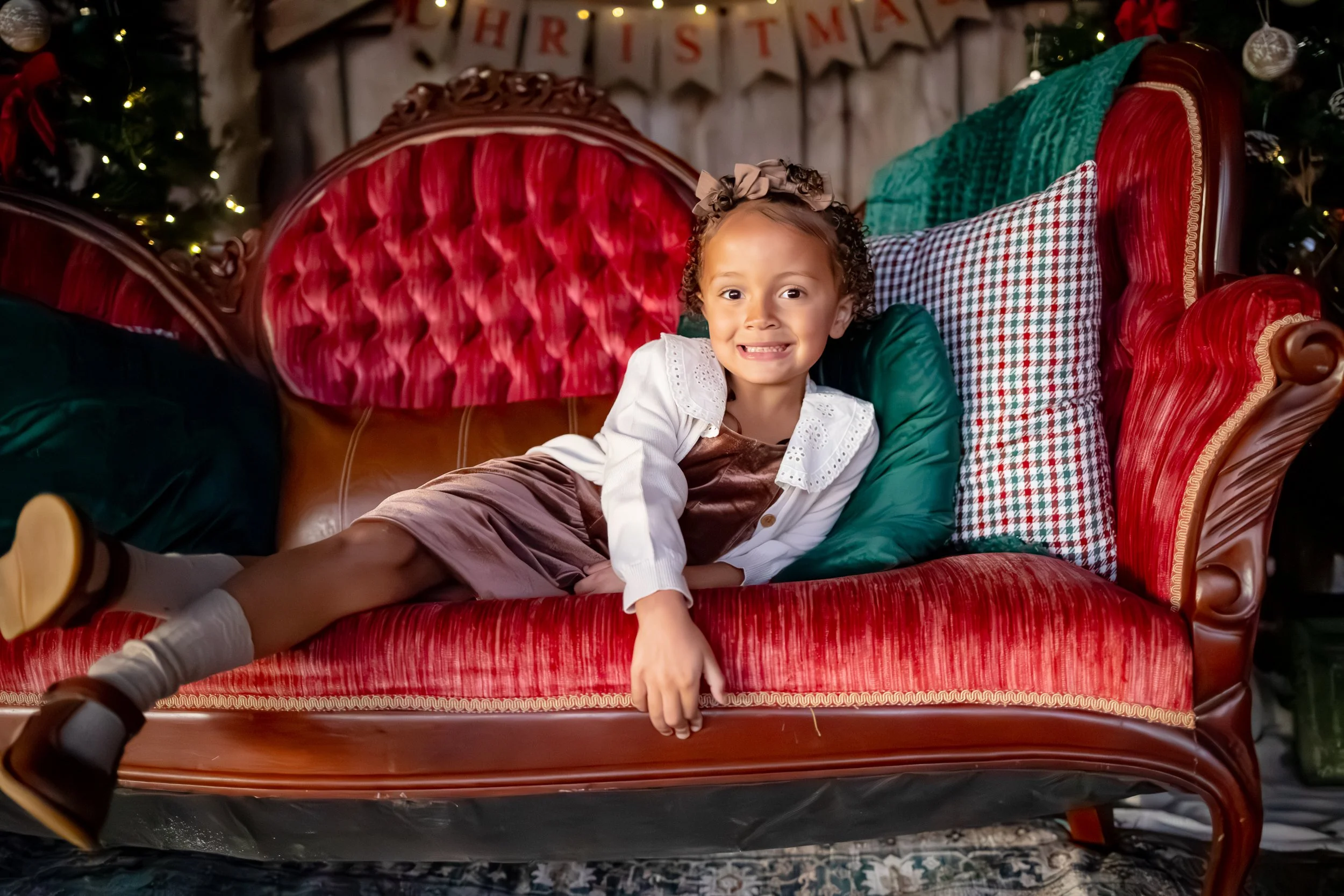 A young girl with curly hair and a bow, smiling while lying on a red vintage sofa with green and checkered pillows. Christmas decorations and a 'Merry Christmas' banner are visible in the background.