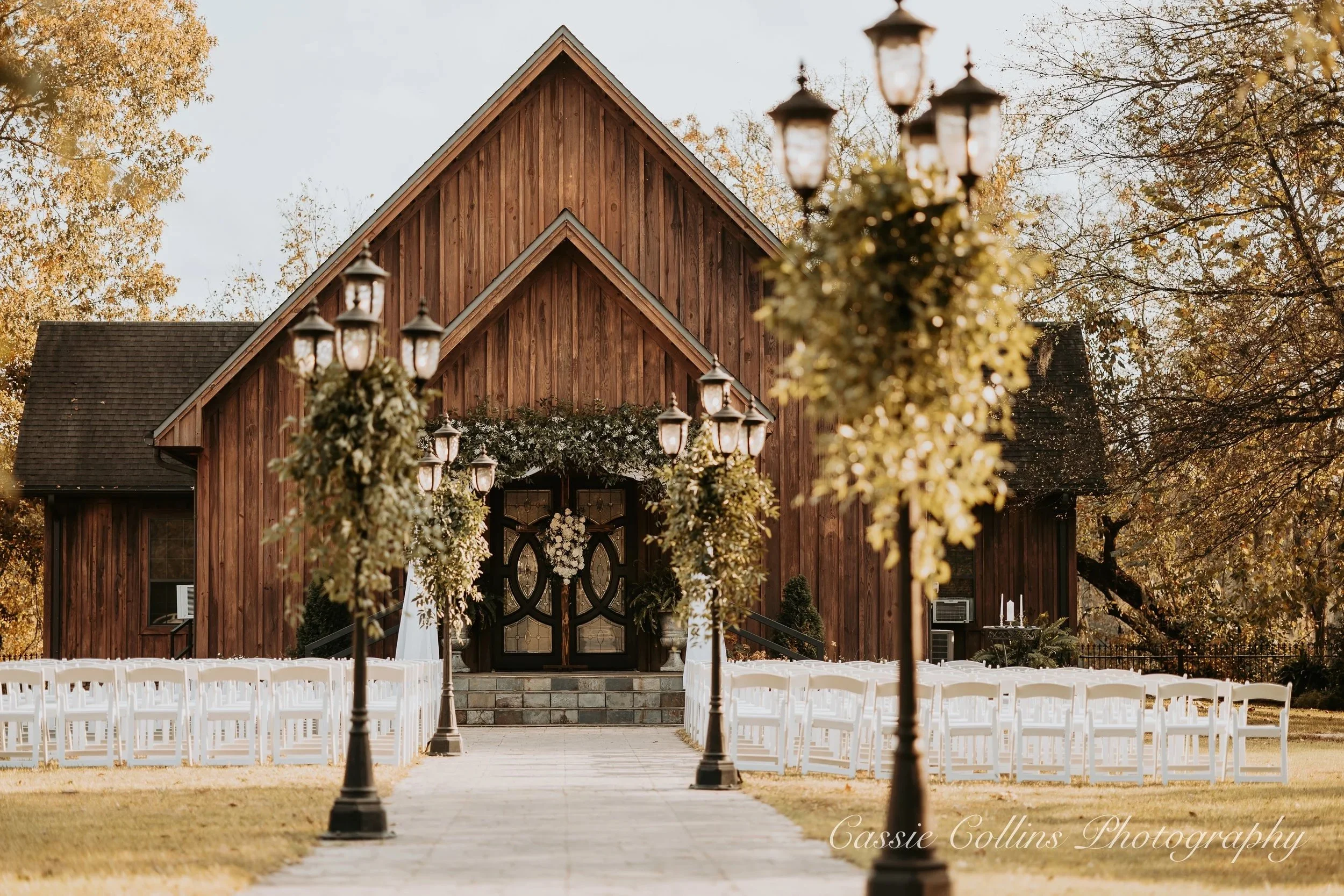 Chapel decorated for a wedding with white chairs, trees, and vintage street lamps leading to the entrance, surrounded by autumn trees.
