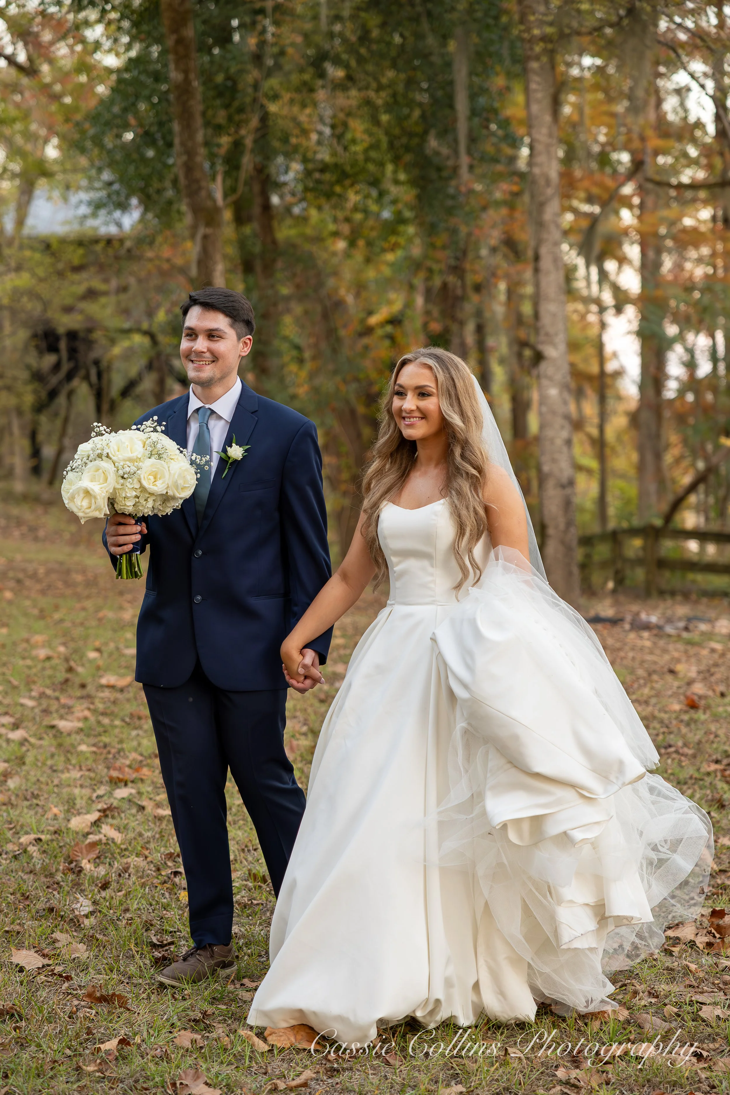 A bride and groom holding hands outdoors in a wooded area during autumn, with the groom holding a bouquet of white roses.