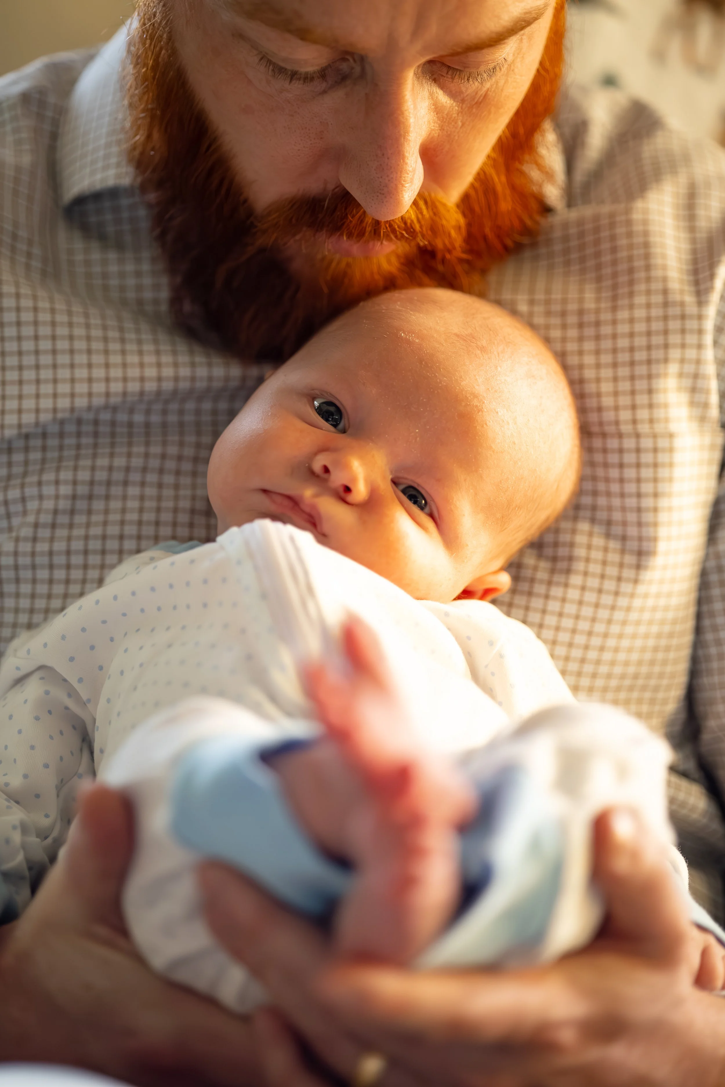 A man with a red beard holding a baby with light skin and big blue eyes, lying on the man's chest, looking at the camera.