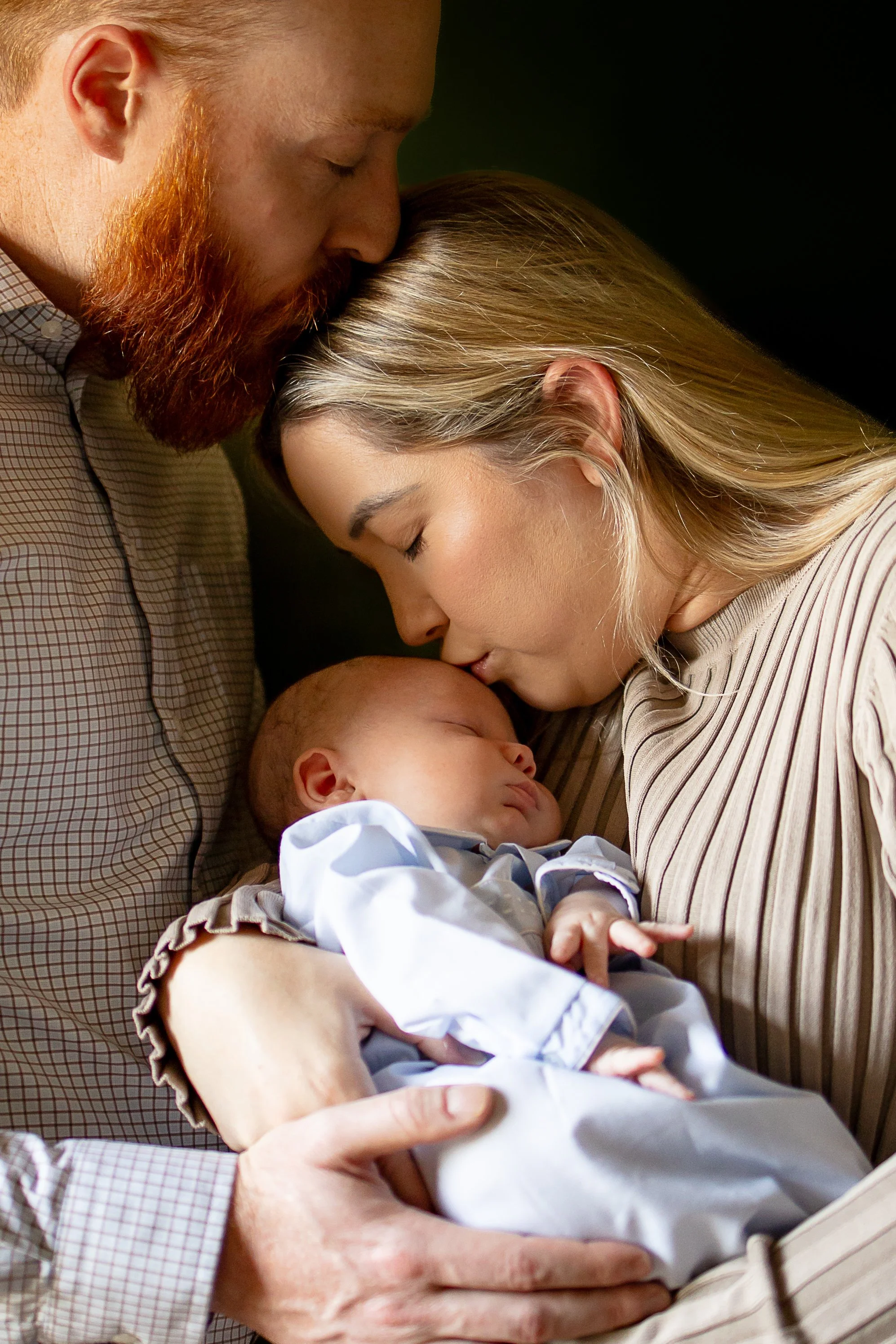 A man and woman gently holding and kissing a sleeping baby.