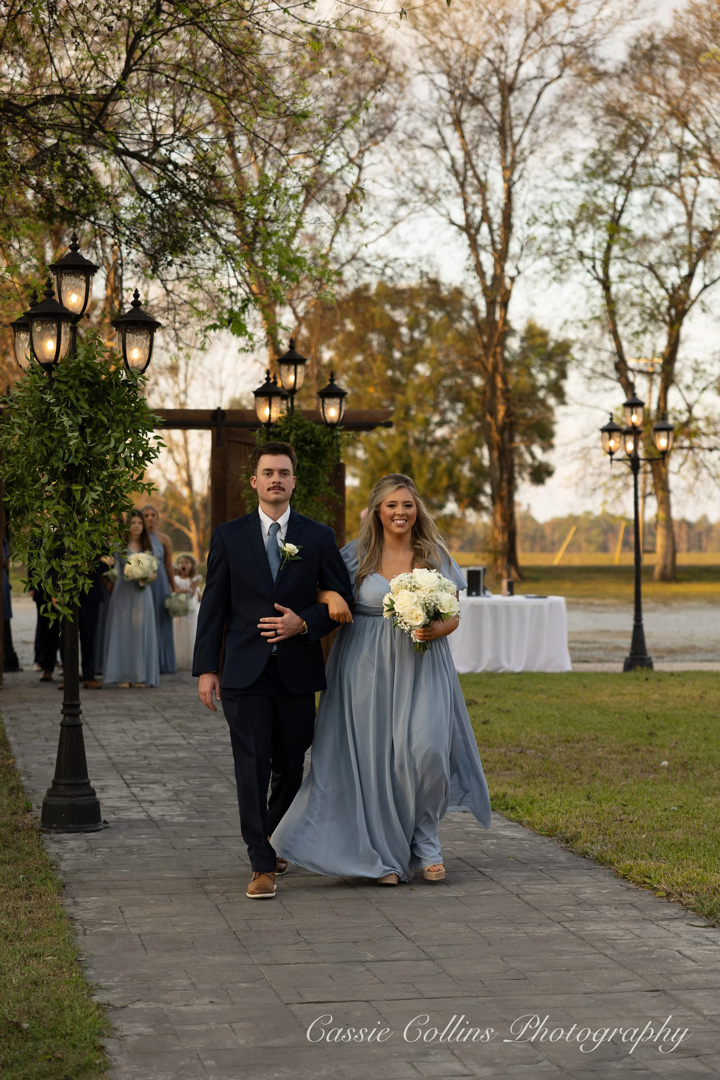 A bride and groom walking down the aisle outdoors during a wedding ceremony at sunset. The bride is holding a bouquet of white flowers, wearing a long, flowing, light blue dress, and has long blond hair. The groom is dressed in a navy suit with a white shirt and matching tie. Bridesmaids and groomsmen are visible in the background, holding bouquets, with trees and lampposts lining the aisle.