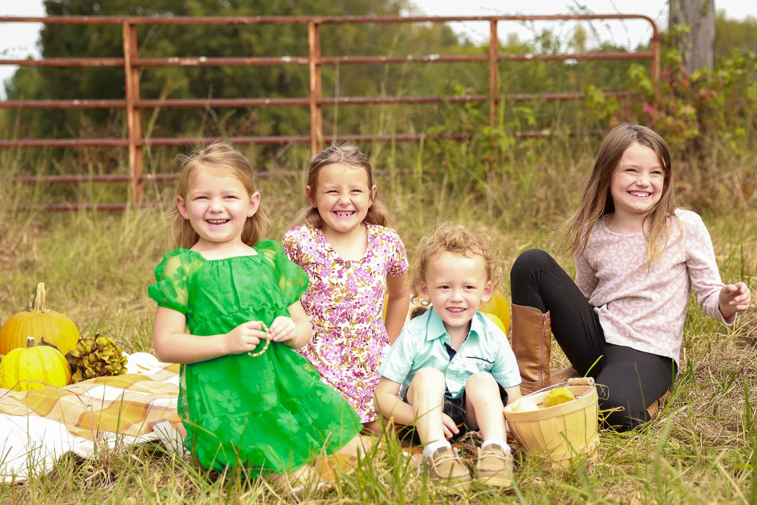 Four children sitting on a picnic blanket in a field, smiling, with pumpkins and autumn decorations around them.
