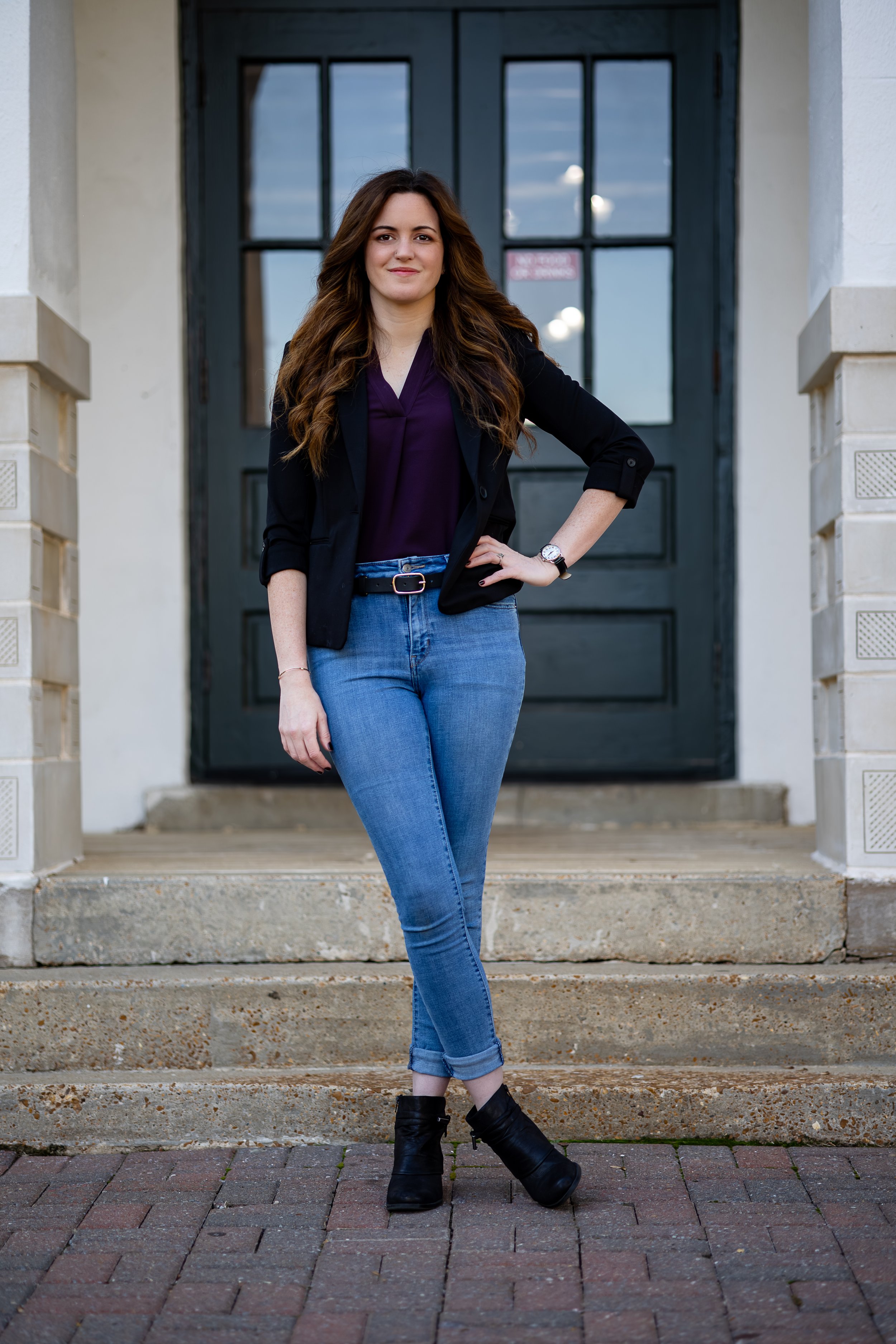 A woman standing on front steps in casual business attire in front of a black door with large glass panes.