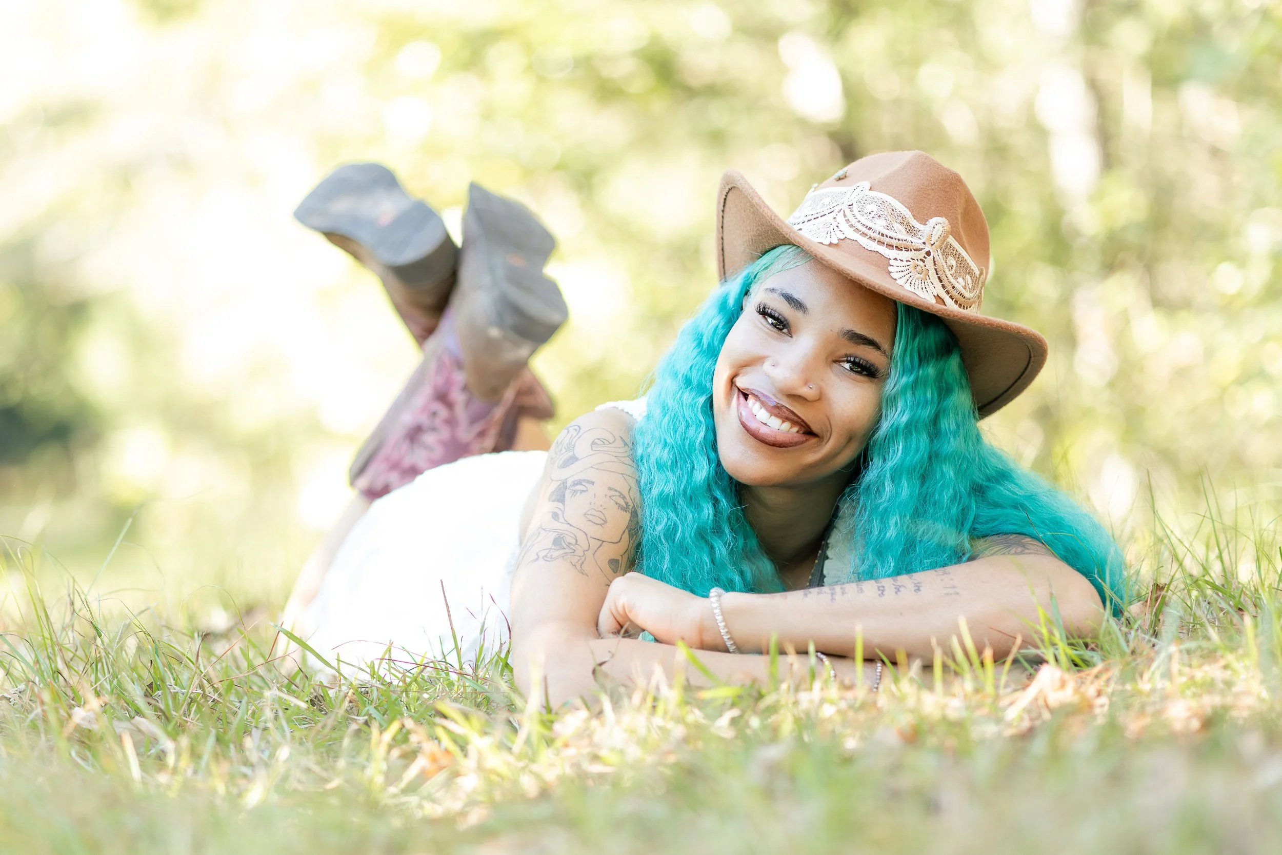 A woman with bright turquoise hair lying on the grass in a park, wearing a tan hat with lace and beige boots, smiling at the camera.