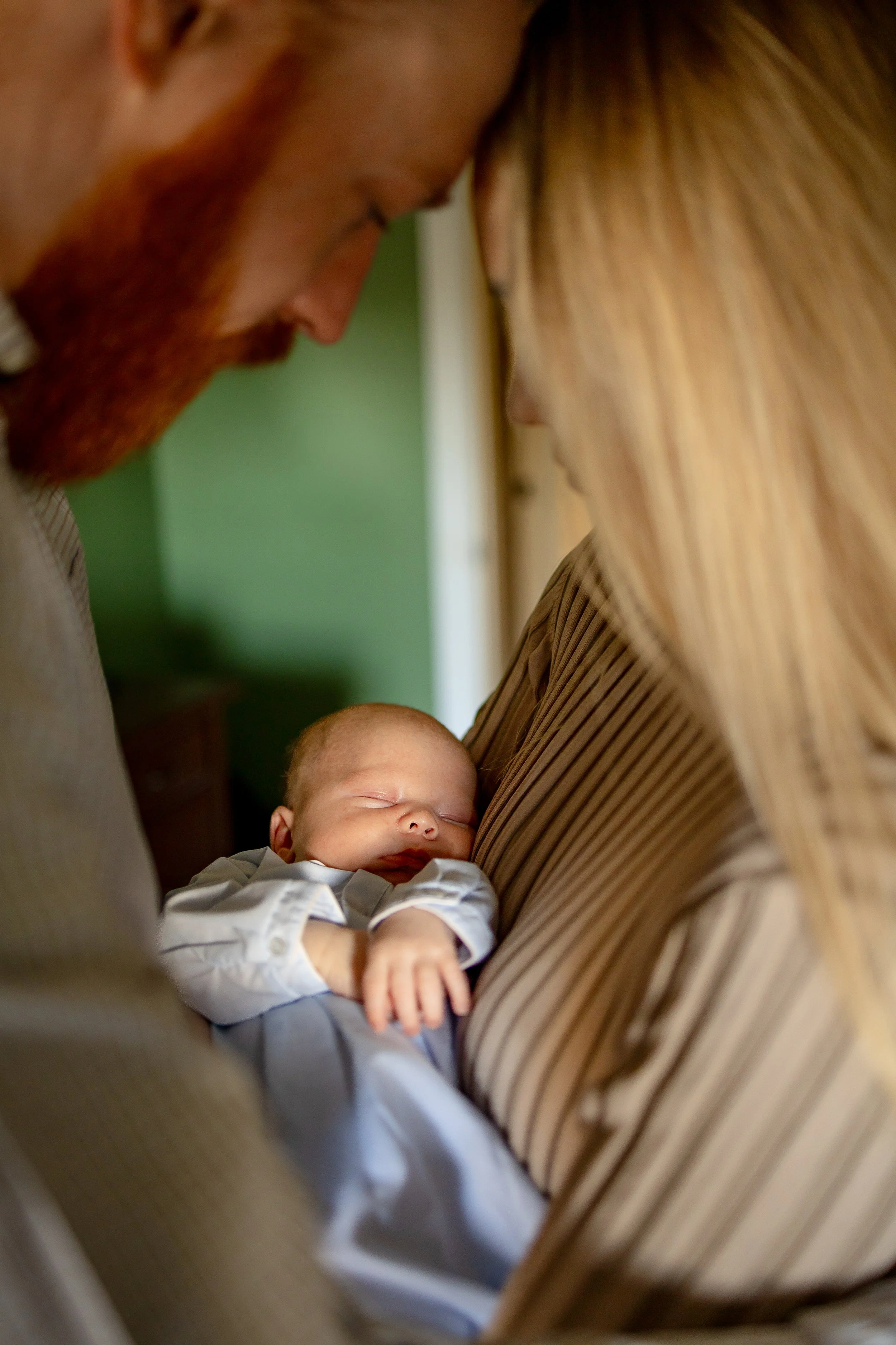 A newborn baby sleeping peacefully in the arms of a woman, with a man leaning close to both of them, all indoors.