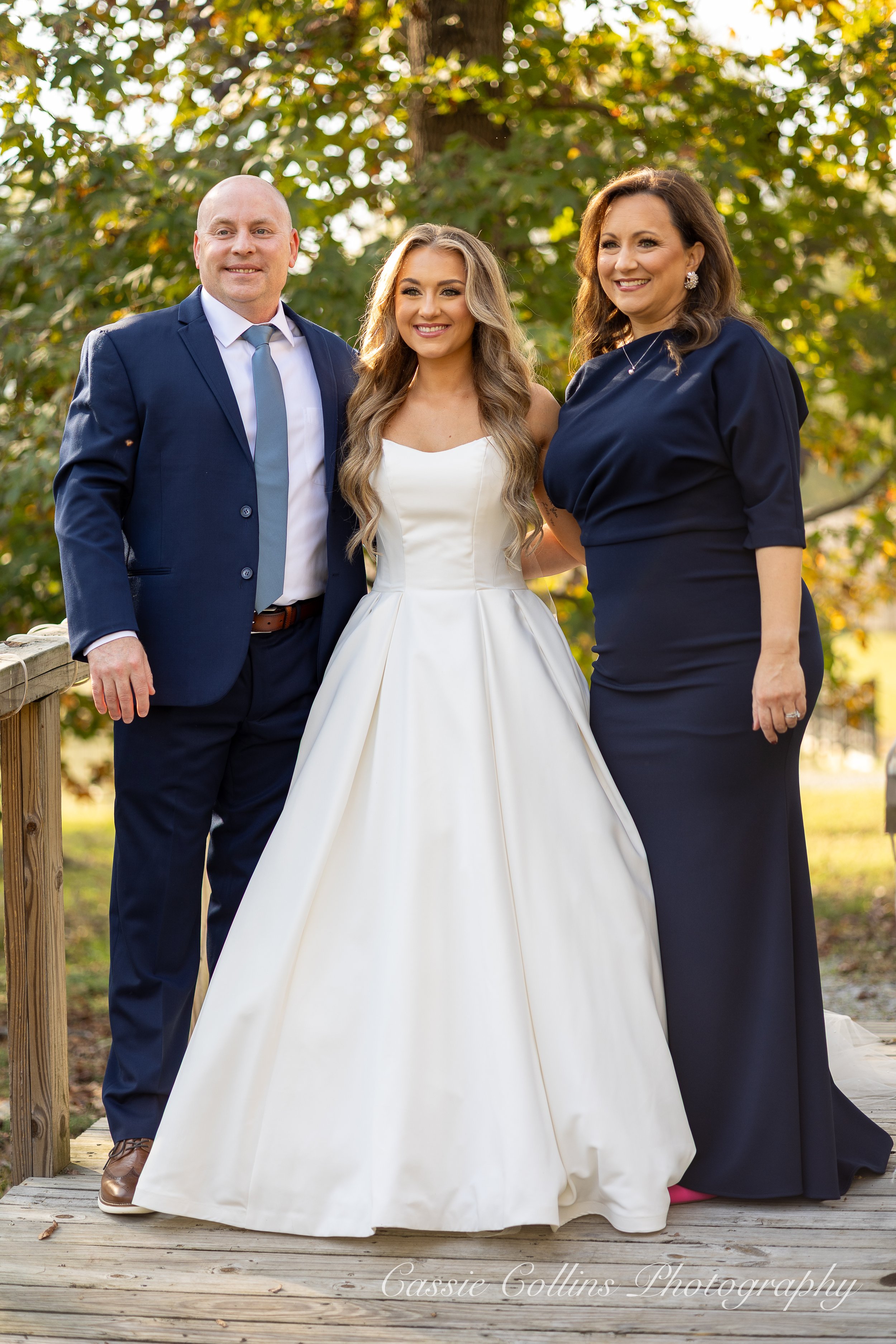 A bride in a white wedding gown standing between a man in a blue suit and a woman in a dark blue dress outdoors on a wooden platform surrounded by trees.