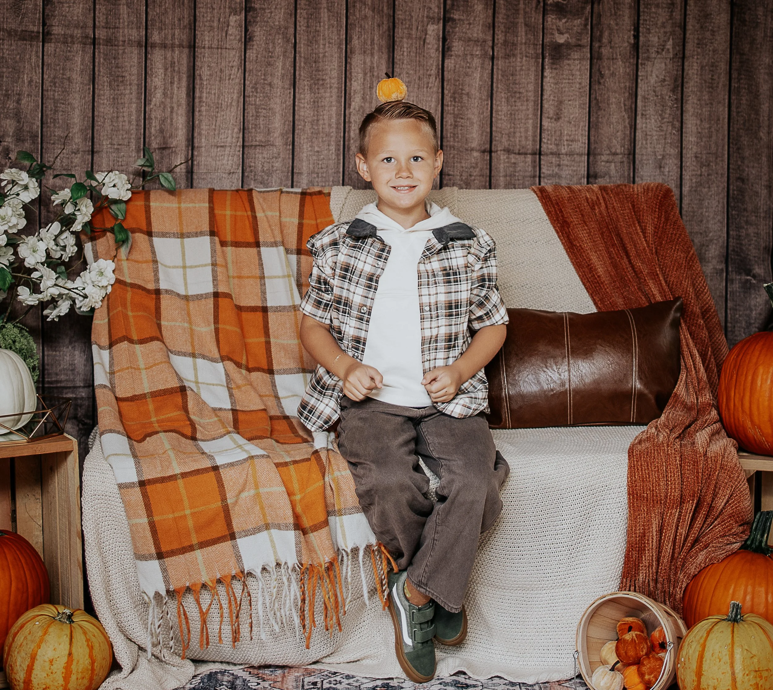 A young boy sitting on a sofa surrounded by pumpkins, with pumpkins also on the sides, a blanket, and fall decor, in front of a wooden wall.