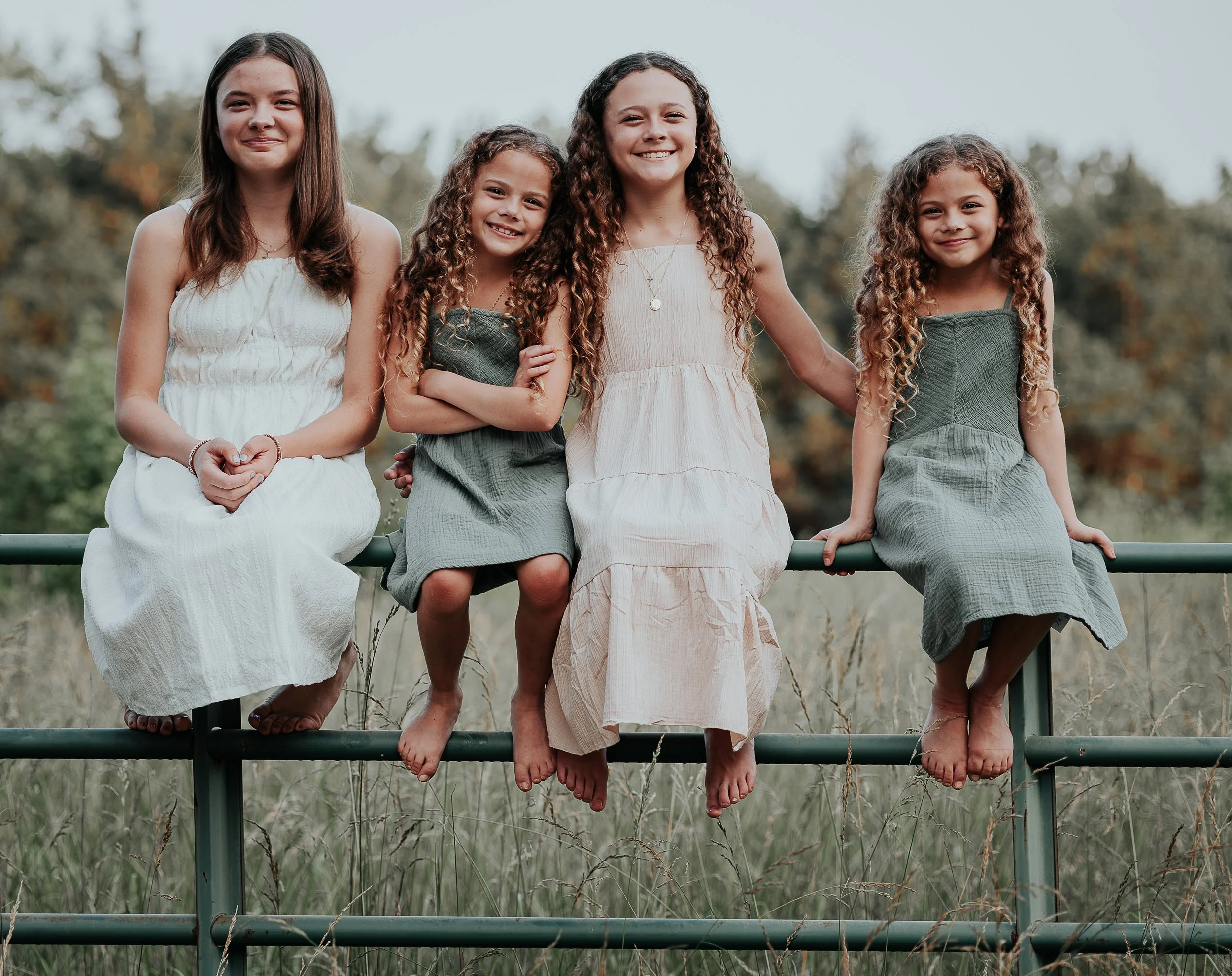 Four young girls sitting on a metal fence outdoors, smiling and looking at the camera, with trees in the background.