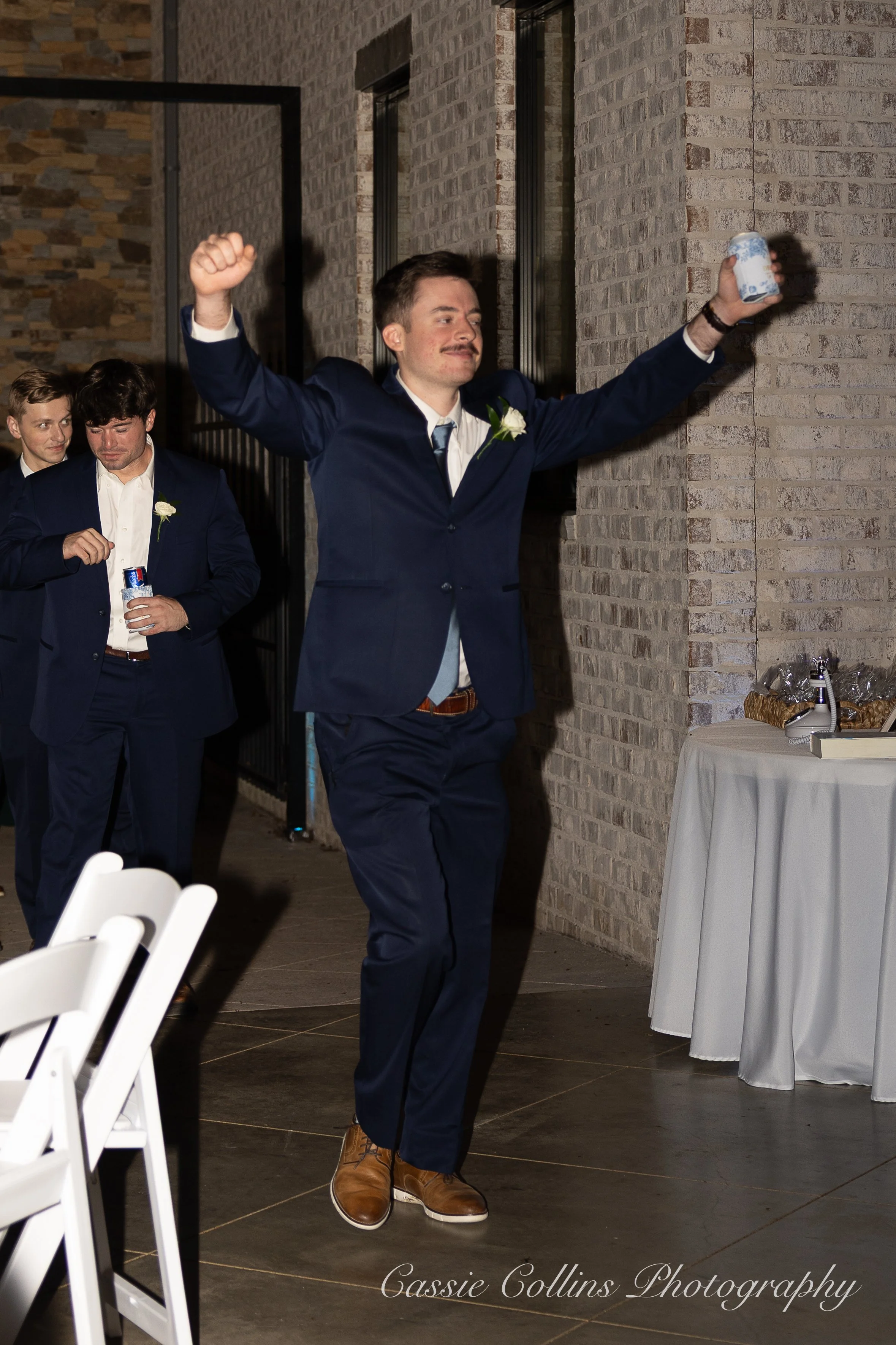 A man in a suit with a boutonniere is holding a can of beverage in one hand and raising his other arm in celebration at a wedding reception, with other men in suits behind him.