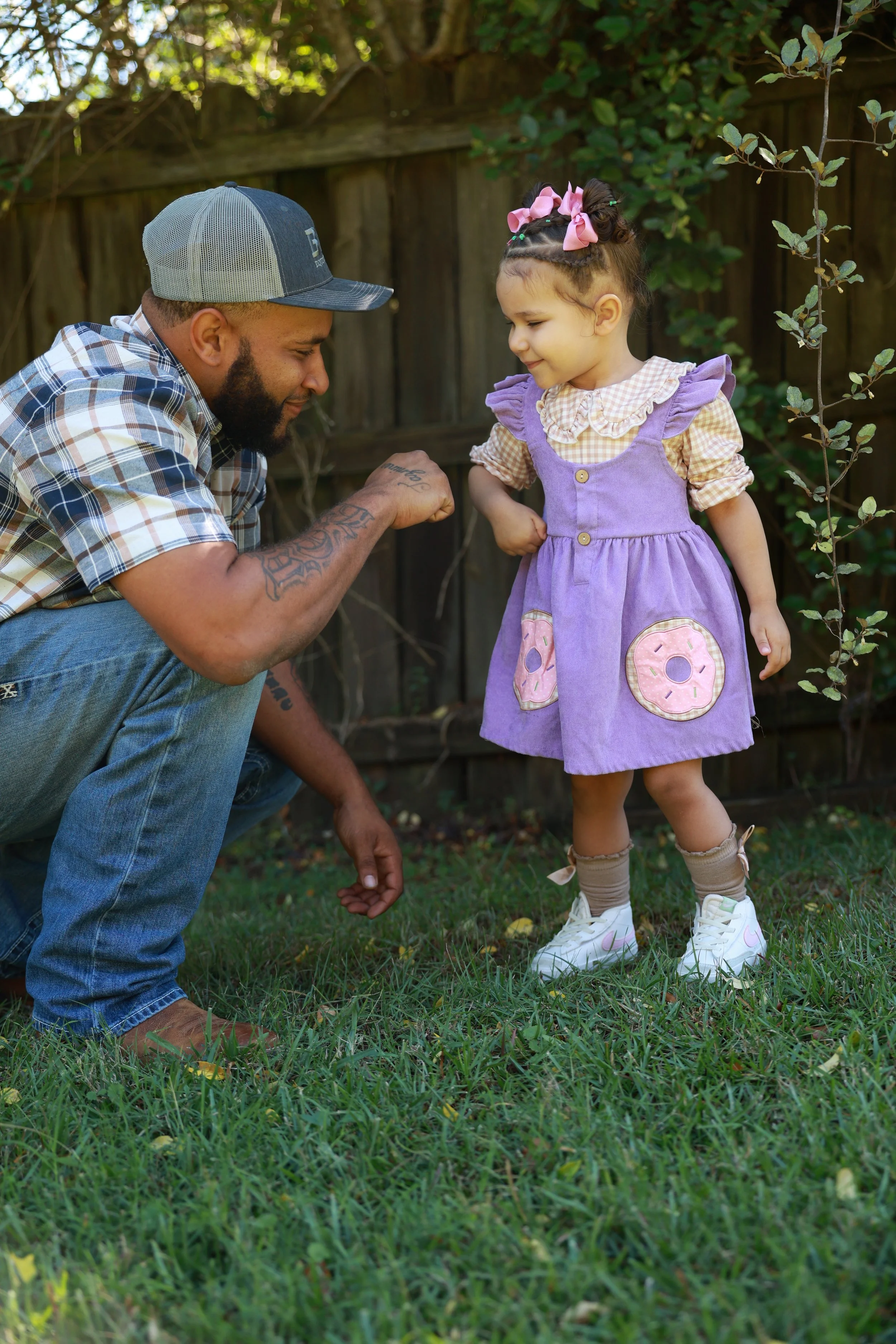 A man in a plaid shirt and baseball cap crouches on the grass, flexing his arm while a young girl in a purple dress with donut patches and pink bows on her head stands in front of him, smiling. They are outdoors in a backyard with a wooden fence and 