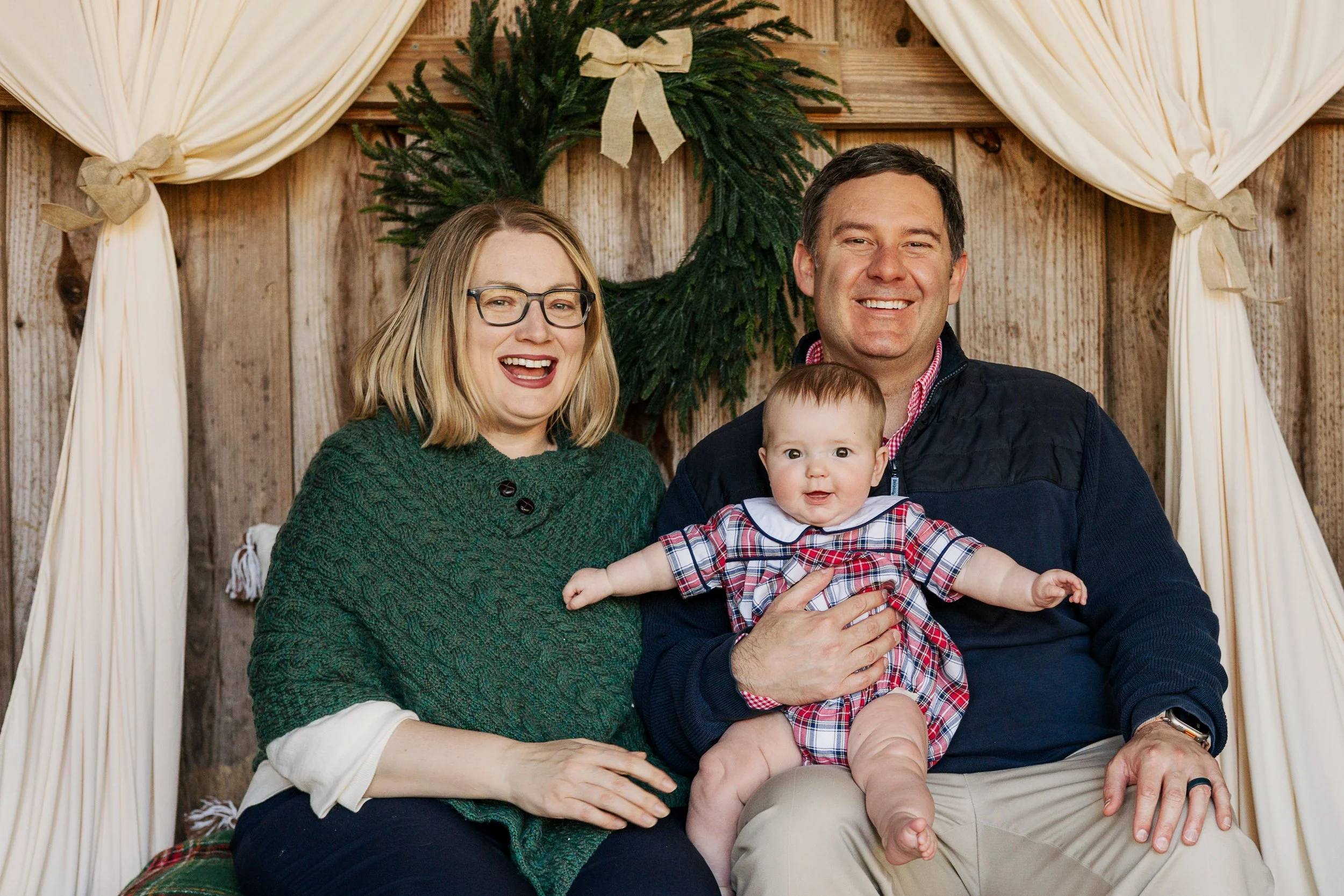 A smiling family of three sitting in front of a wooden wall with a Christmas wreath, with cream curtains tied back, during a holiday celebration.