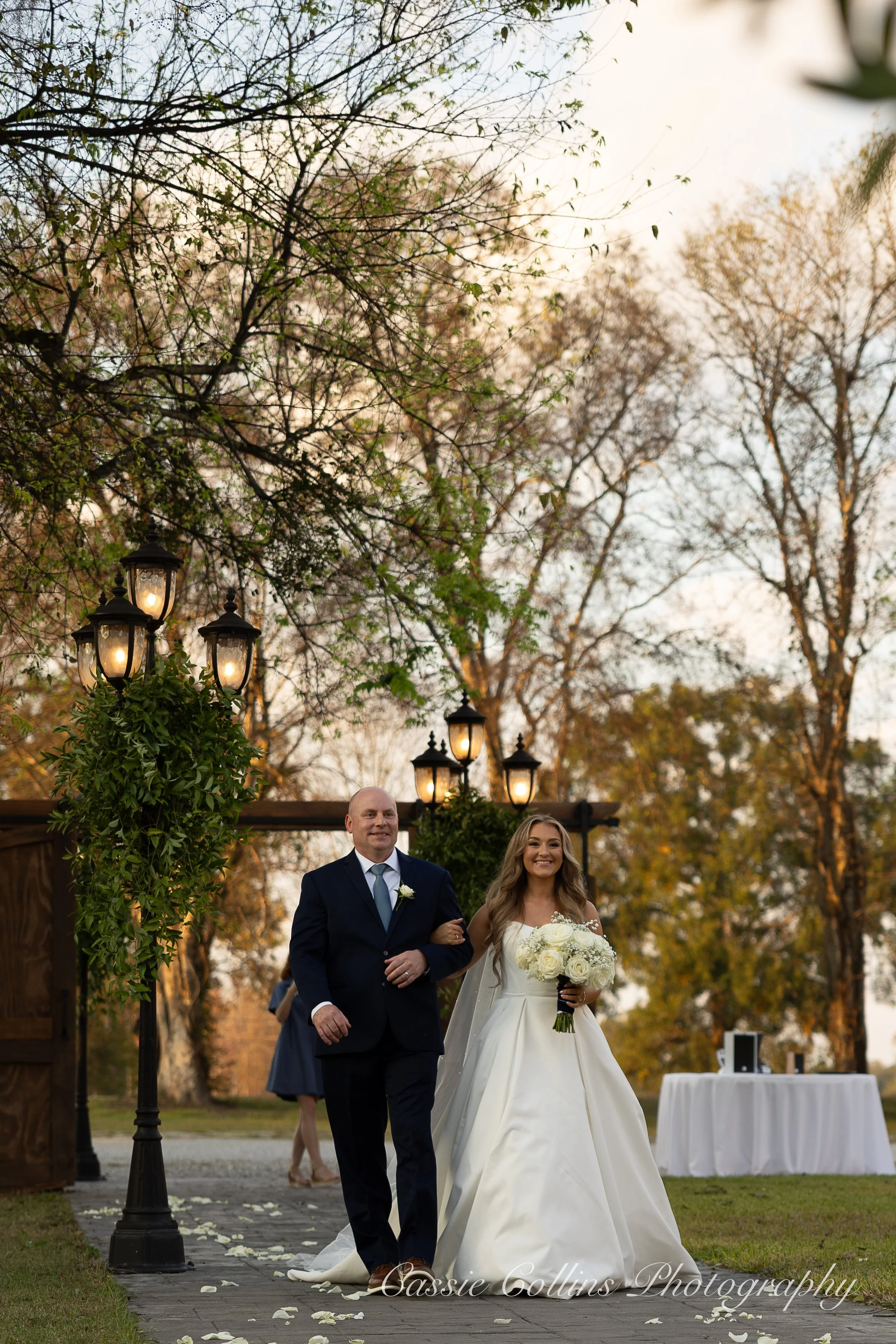 Bride walking arm-in-arm with a man during an outdoor wedding ceremony at sunset, with lanterns and trees in the background.