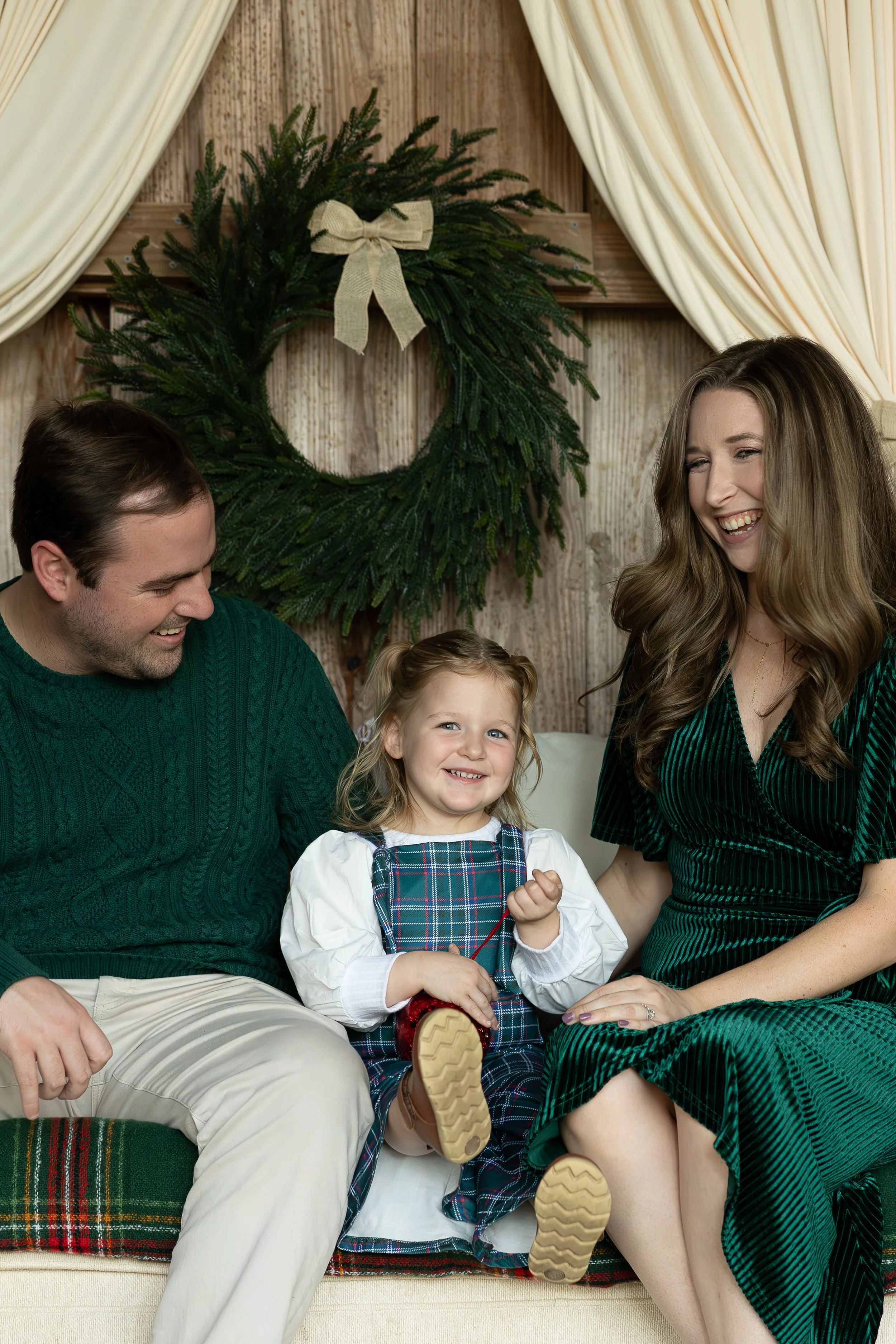 Family of three, two adults and a young girl, sitting together on a couch during Christmas, with a wreath hanging on a wooden wall behind them and cream curtains framing the scene.