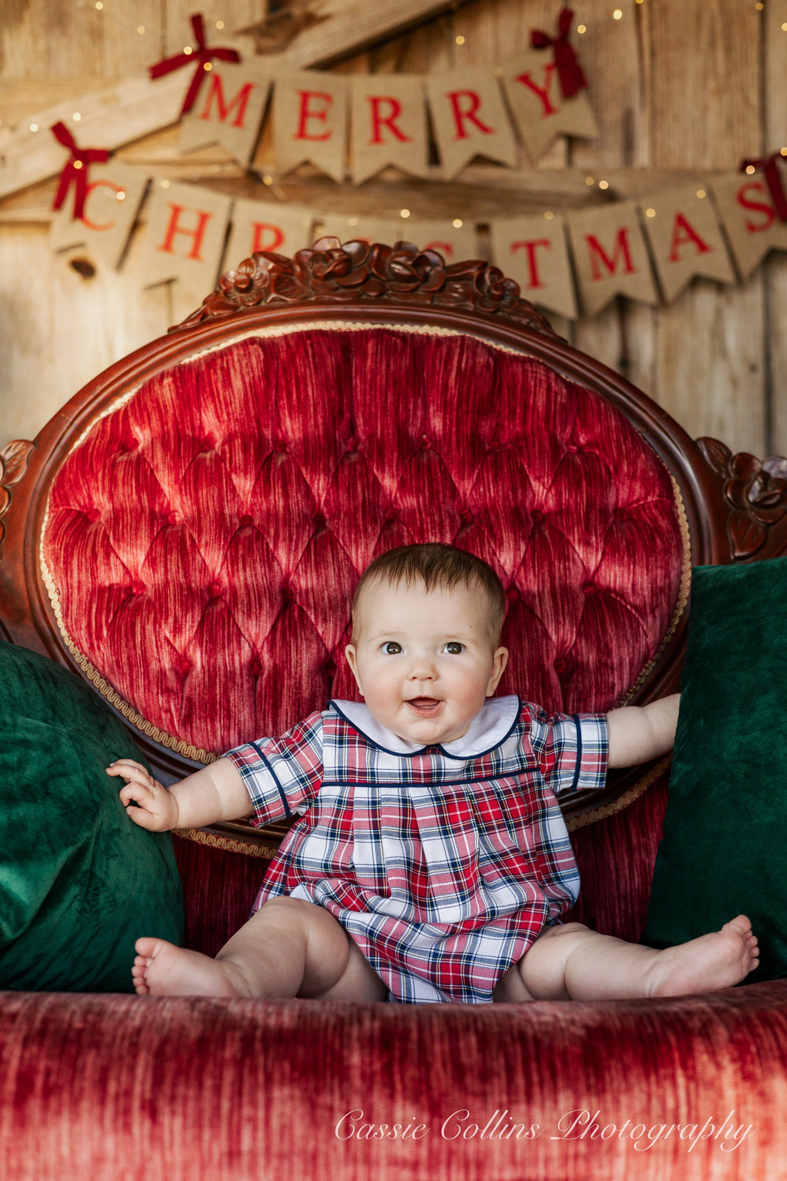 A baby sitting on a red velvet antique sofa, with Christmas decorations and a "Merry Christmas" banner in the background.