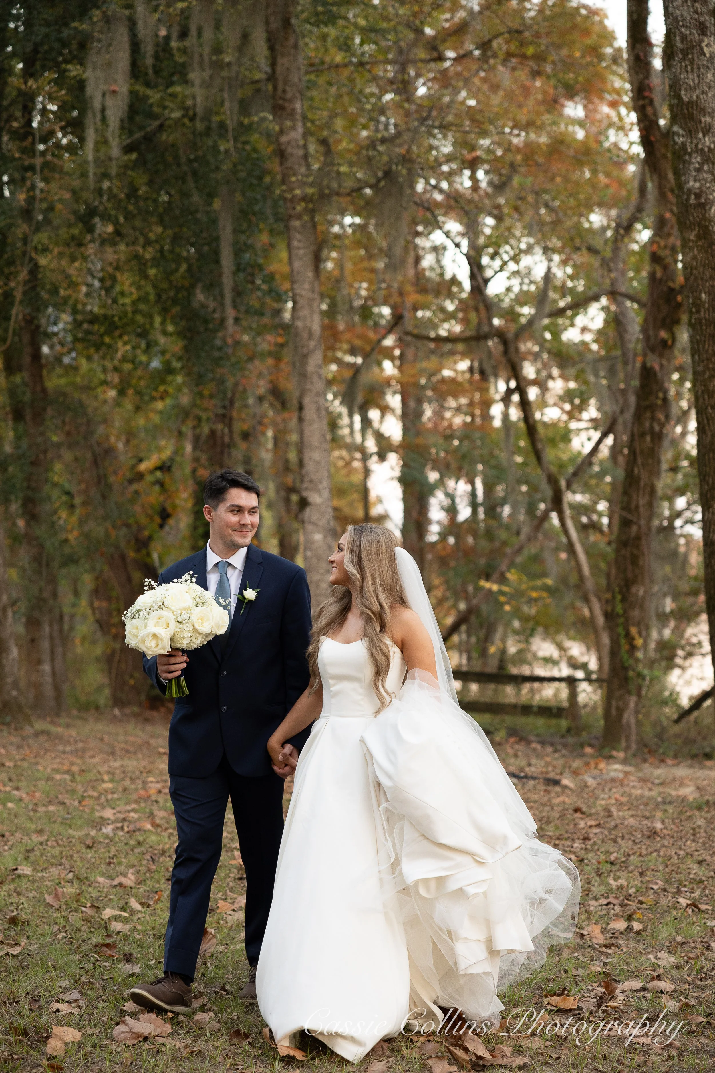 A bride and groom walking hand in hand through a wooded outdoor setting during their wedding. The bride is wearing a white wedding dress and veil, holding her dress with one hand. The groom is dressed in a navy suit holding a bouquet of white flowers, smiling at her.