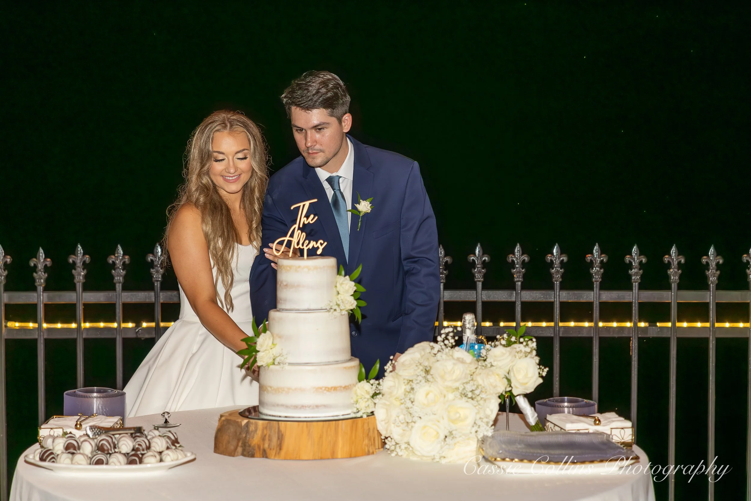 A bride and groom cut a wedding cake outdoors at night, with the bride smiling and the groom looking serious, a wedding cake with white icing and flowers, a bouquet of white roses, and a table with chocolates and favors.