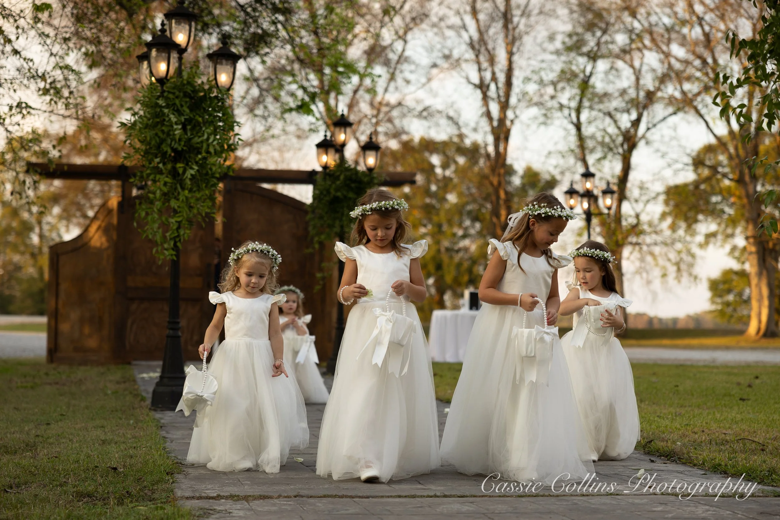 Five young girls in white dresses and flower crowns walking outdoors during a wedding or special event, with lanterns and trees in the background.