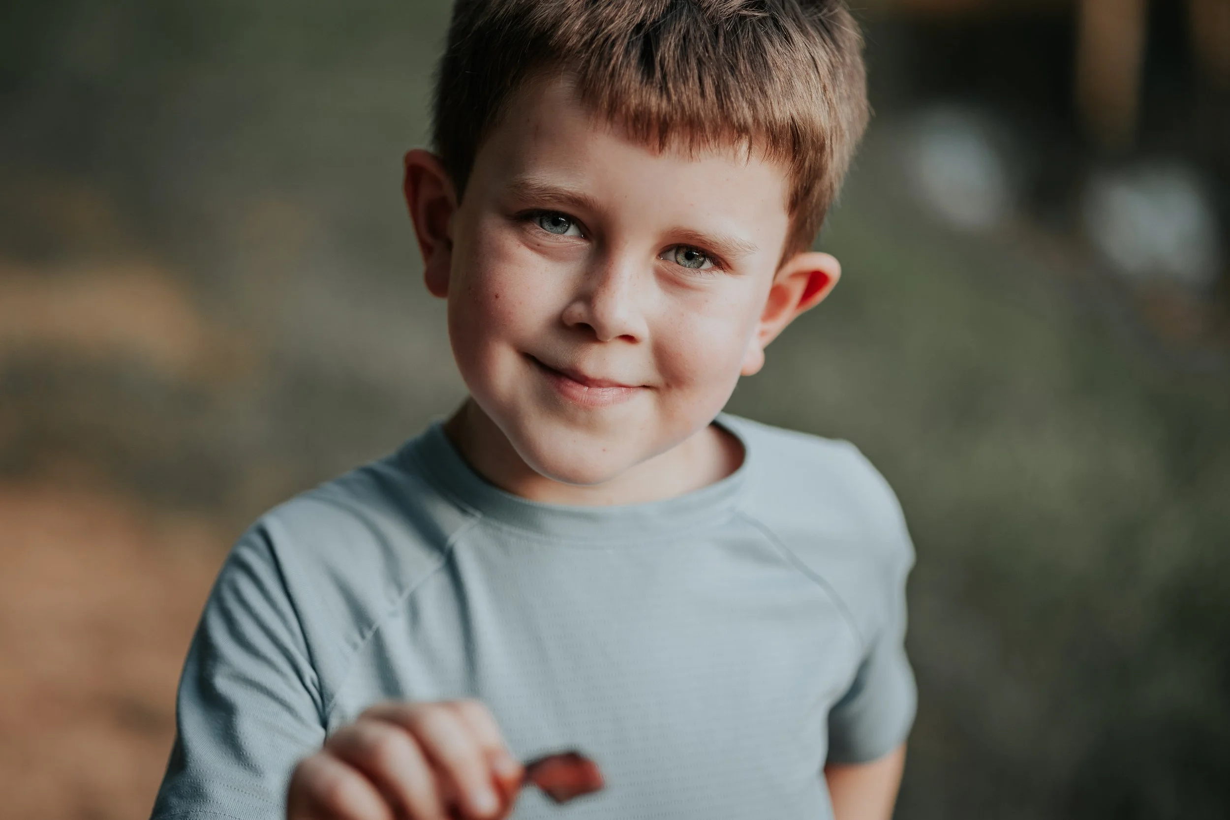 A young boy with short brown hair and blue eyes smiling outdoors, holding a small object in his hand, with a blurred natural background.