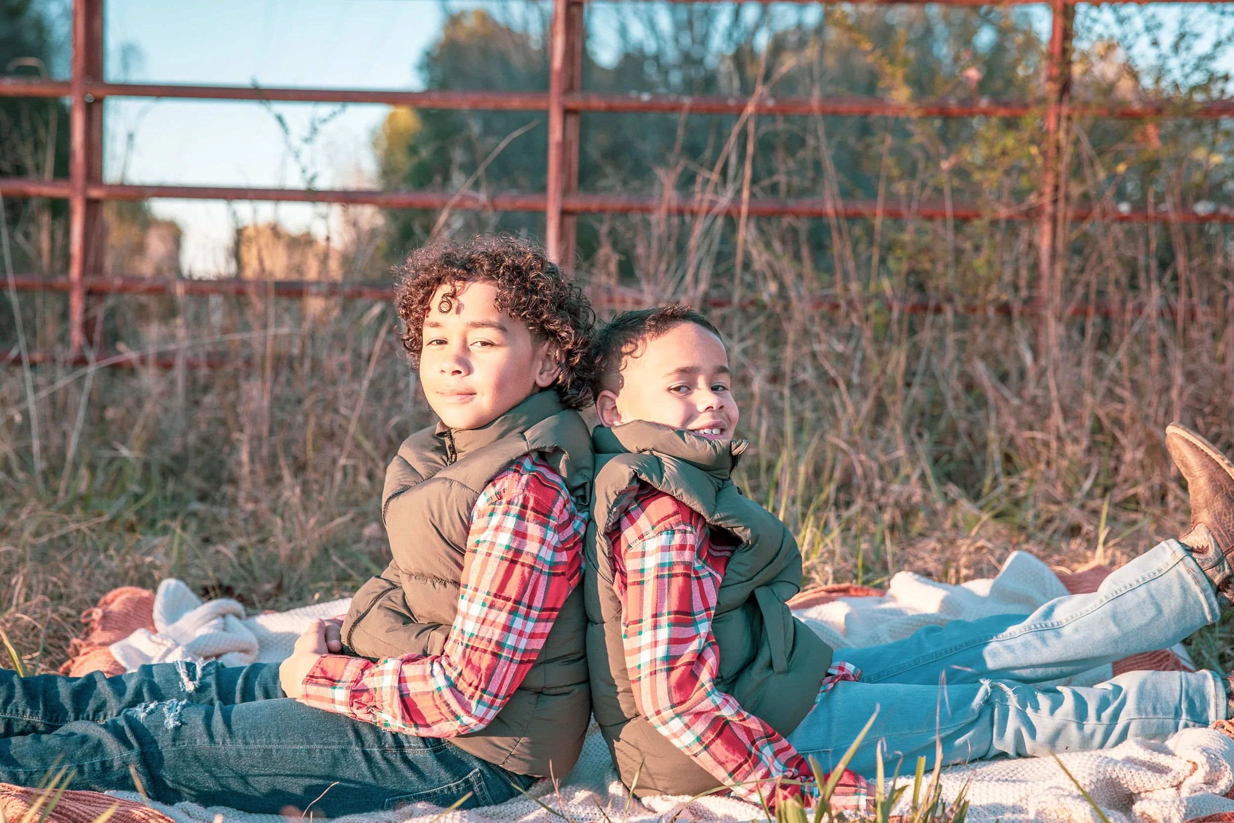 Two children sitting back-to-back on a blanket outdoors, smiling and relaxing in a natural setting with tall grass and a rusty metal fence in the background.