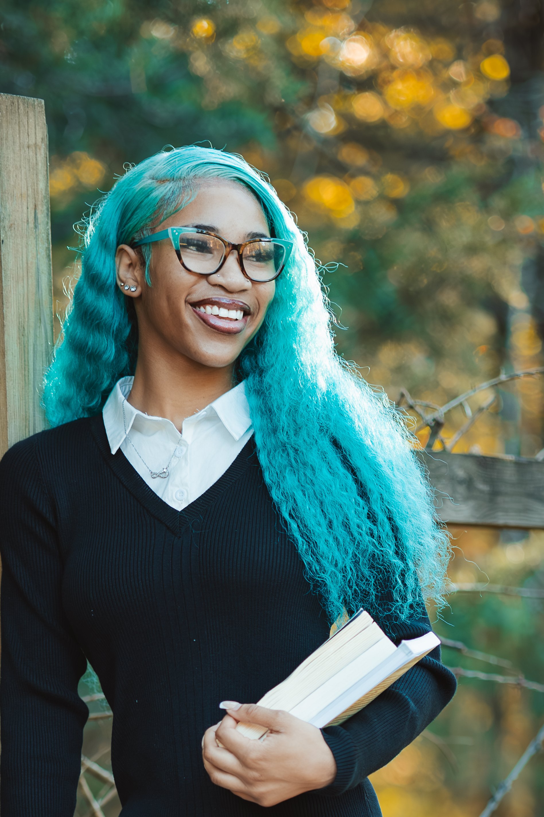 Smiling woman with long blue curly hair and glasses, holding a book, outdoors with blurred autumn trees in background.