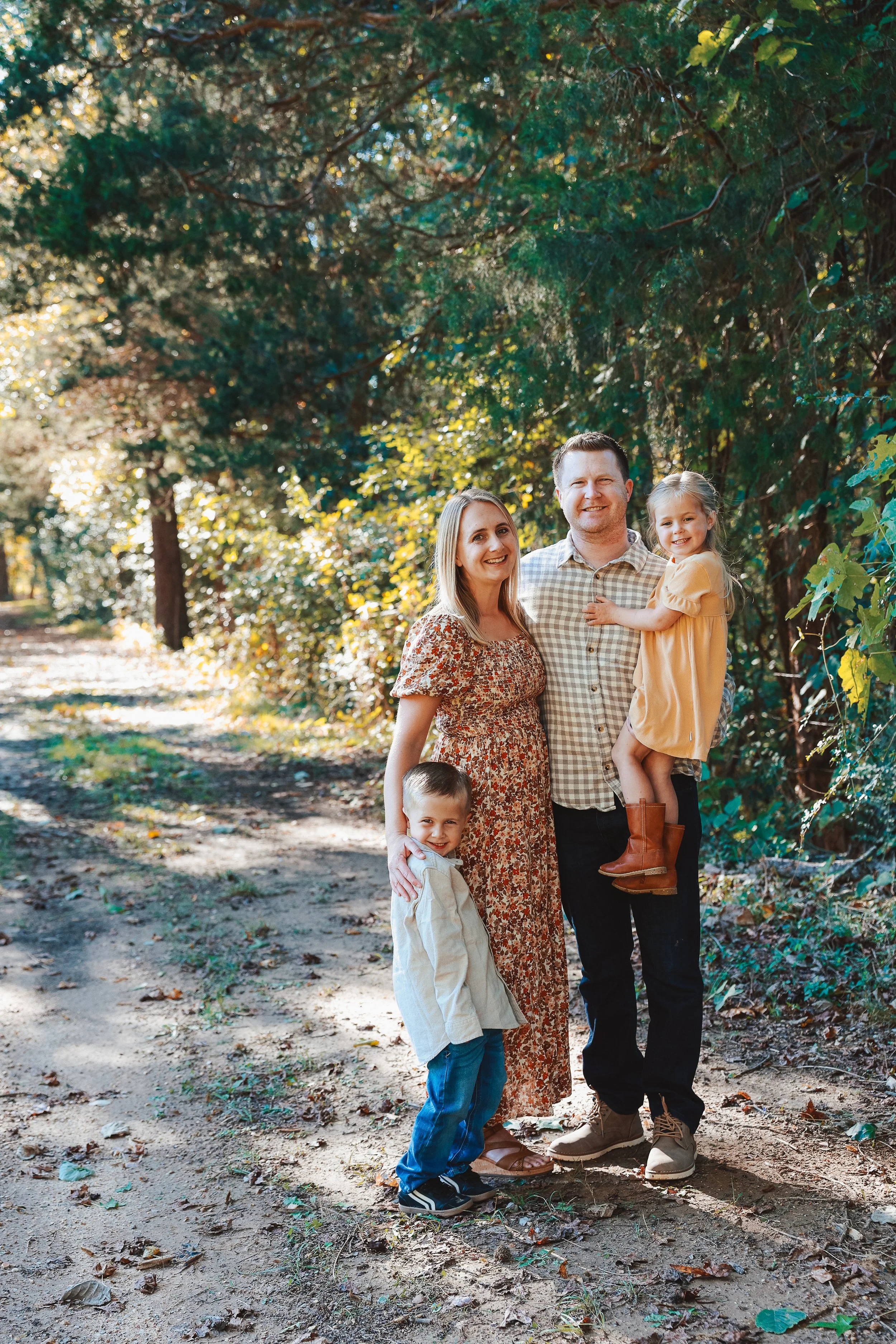 A family of four standing on a trail in a wooded park during daytime. The mother, father, daughter, and son are smiling and together in nature.