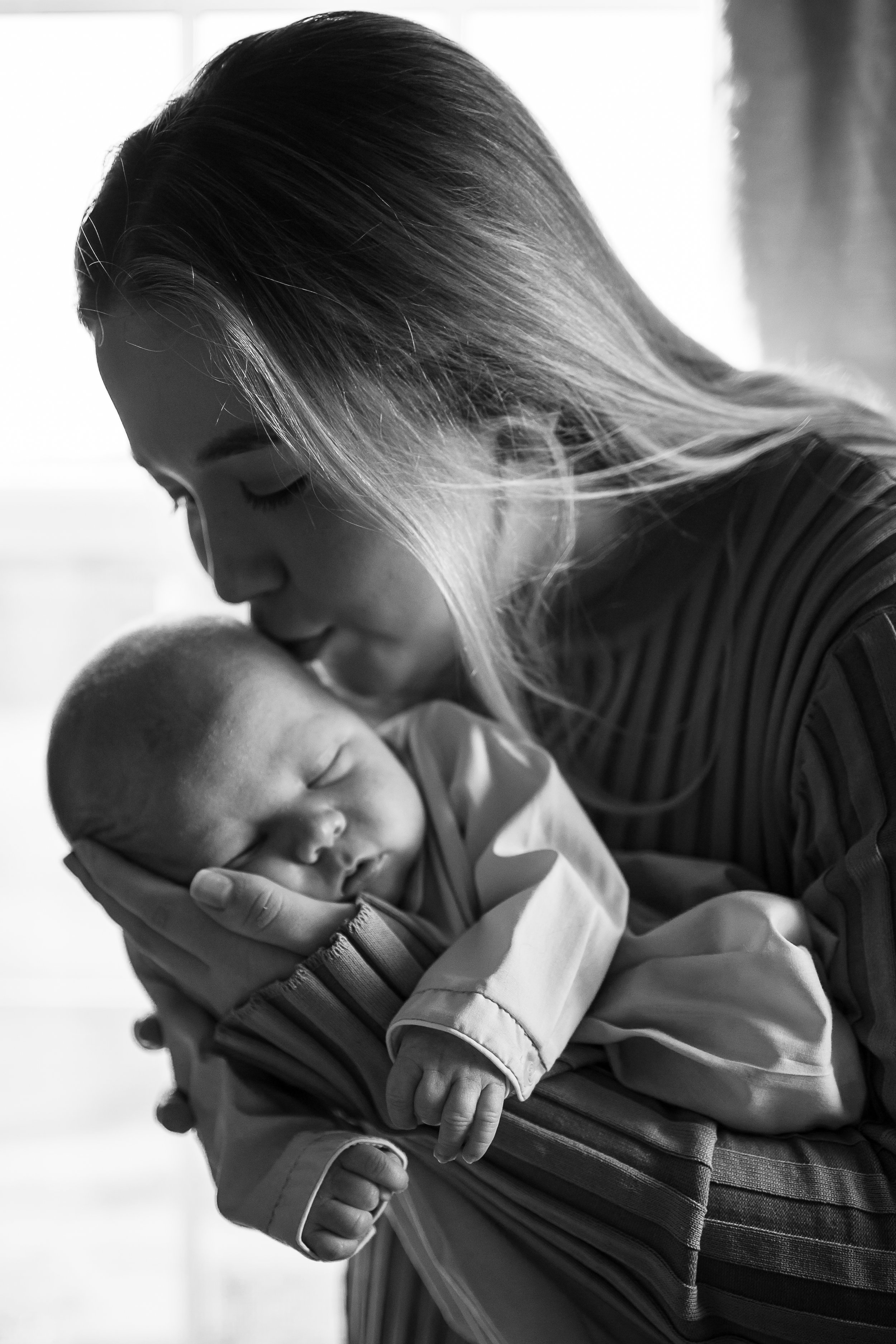 A woman tenderly holding a sleeping baby close to her face, with the woman's eyes closed and her lips touching the baby's forehead in an intimate moment.