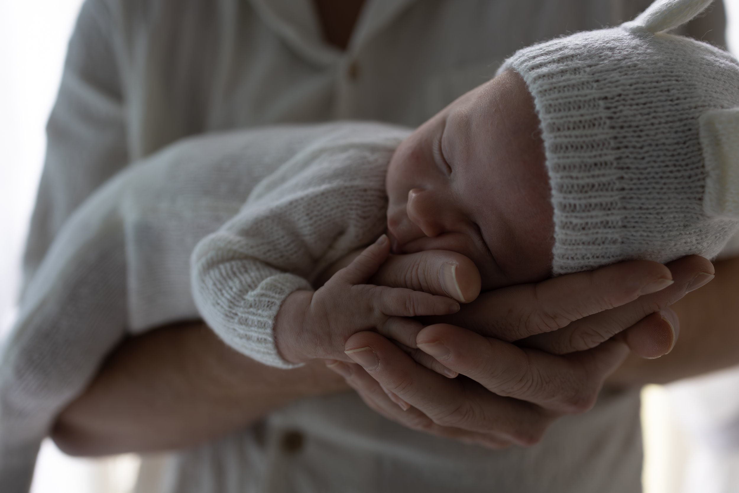 Close-up of a newborn baby, wearing a knit hat and sweater, peacefully sleeping in an adult’s hands.