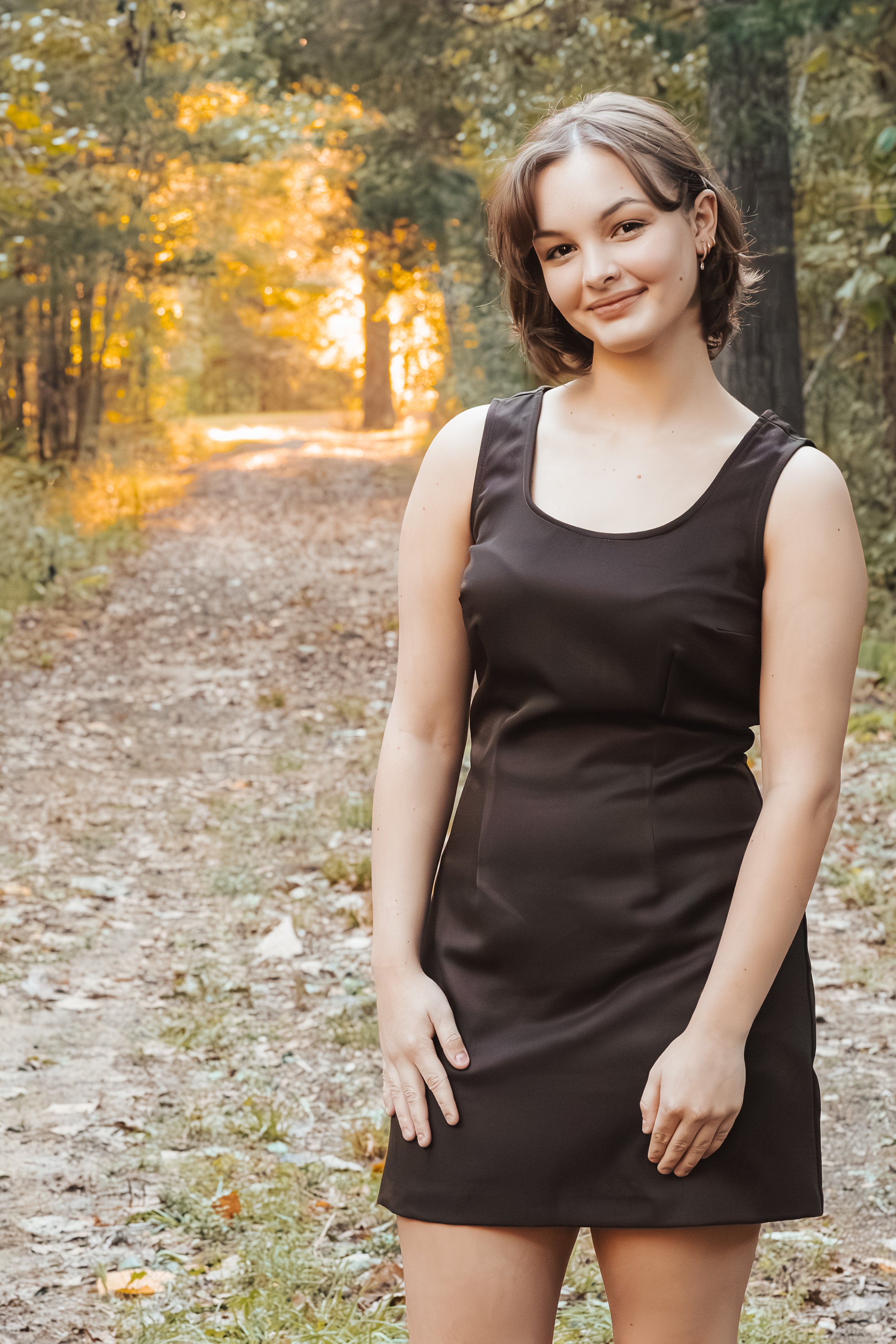 A young woman with short brown hair and earrings outdoors, wearing a black sleeveless dress, standing on a wooded trail during sunset.