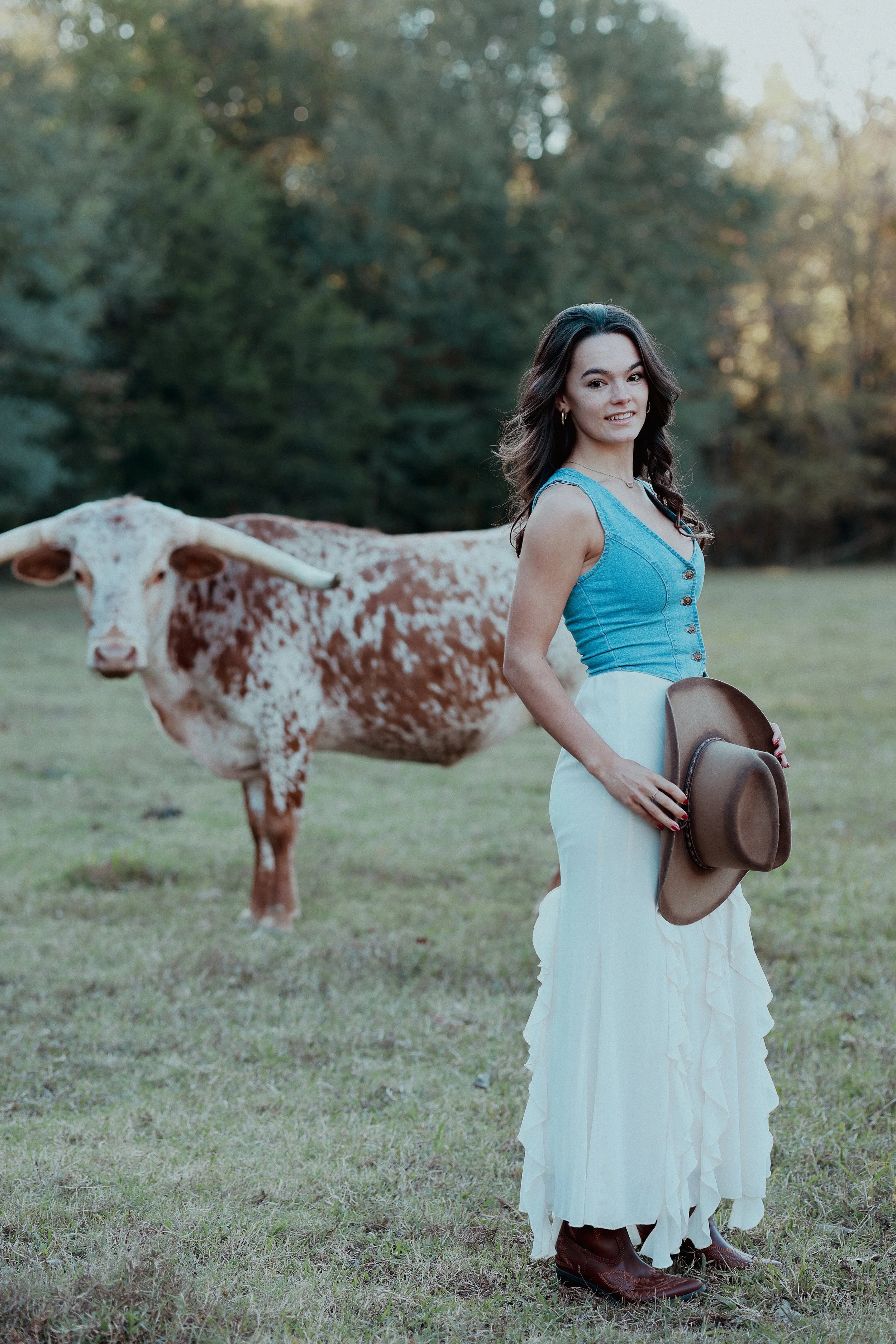 A woman in a blue sleeveless top, white long skirt, brown cowboy boots, and holding a cowboy hat, standing on a grassy field with a brown and white speckled cow in the background, trees, and late afternoon sunlight.