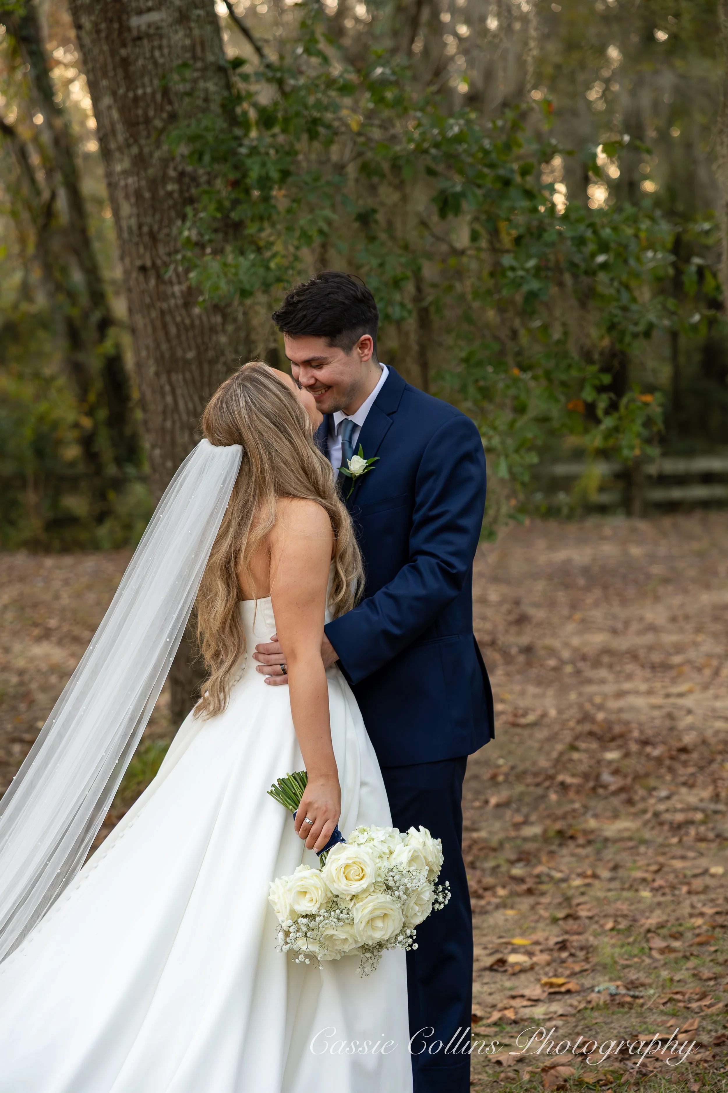 A bride and groom sharing a kiss outdoors during their wedding, with the bride holding a bouquet of white roses and the groom wearing a navy blue suit and tie, in front of trees and nature.