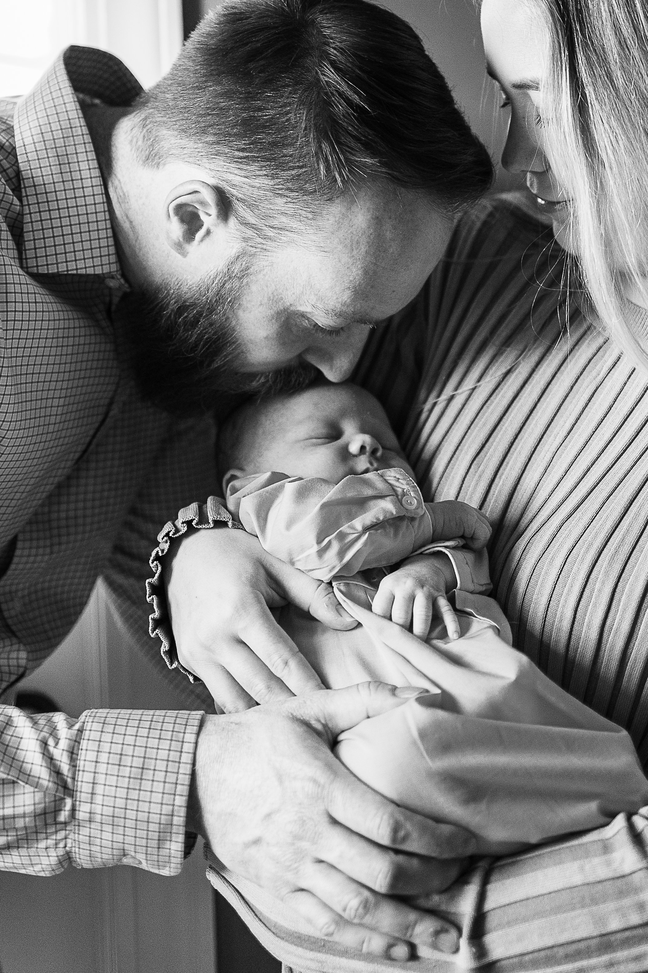 A black and white photo of a man and woman holding a sleeping baby, with the man gently kissing the baby's head.