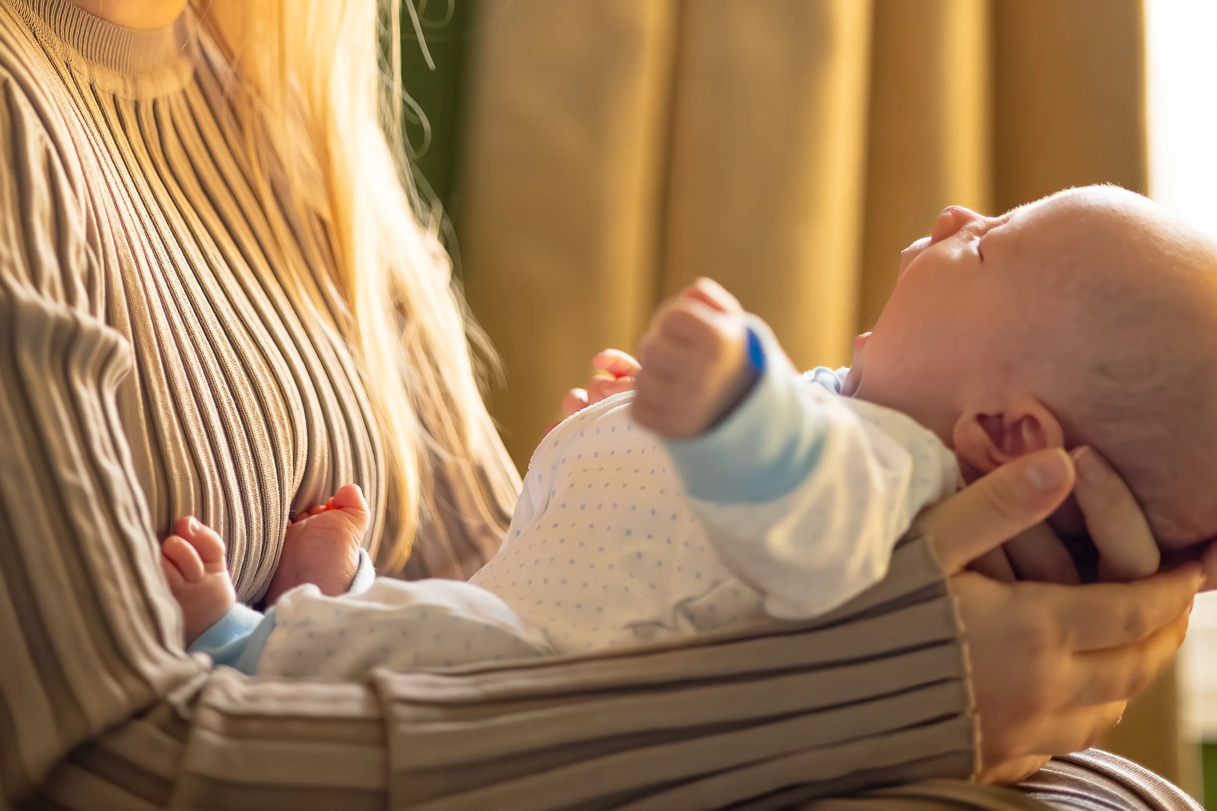 A person holding a sleeping newborn baby in their arms, with the baby lying on their chest. The scene appears to be indoors, with warm lighting and curtains in the background.