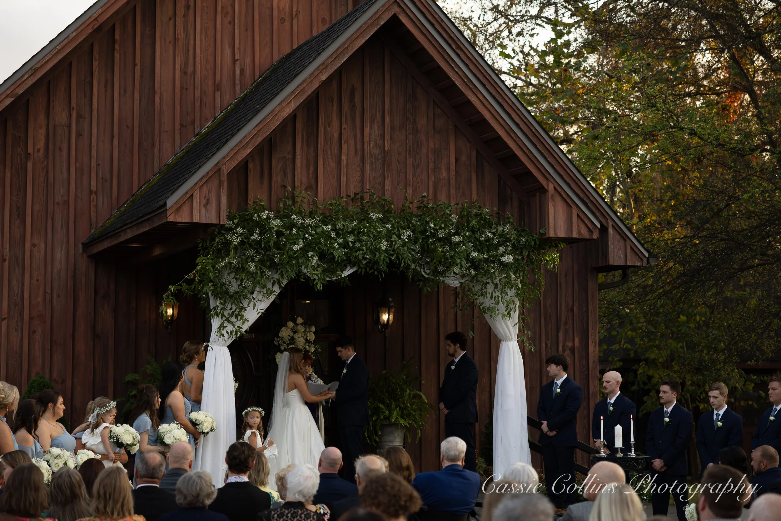 A wedding ceremony taking place outdoors on a wooden stage decorated with greenery and white curtains, with the bride and groom exchanging vows surrounded by bridesmaids and groomsmen, and an audience watching.