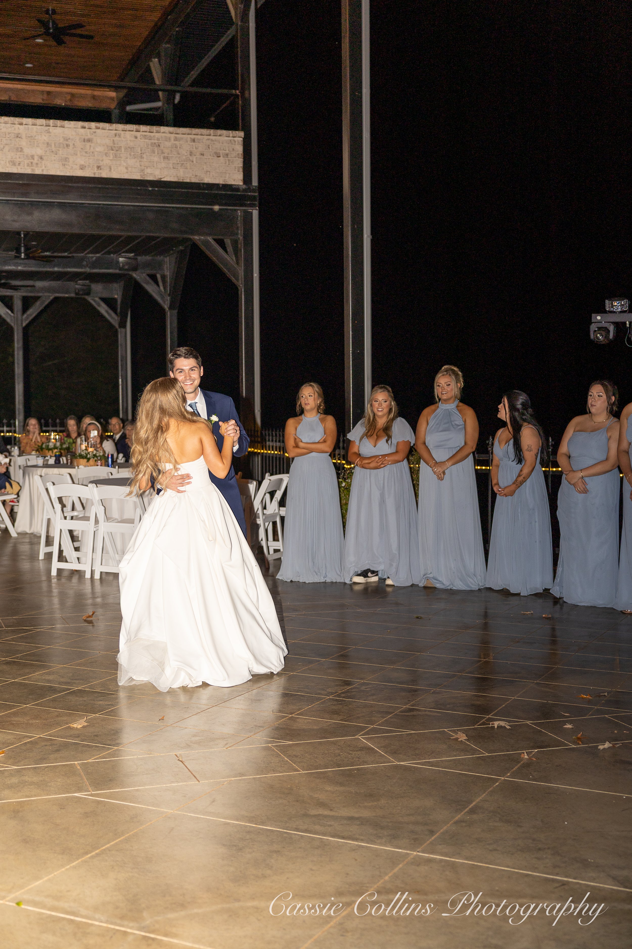 A bride and groom dancing at their wedding reception, with bridesmaids in matching blue dresses standing in the background on a dark outdoor venue.