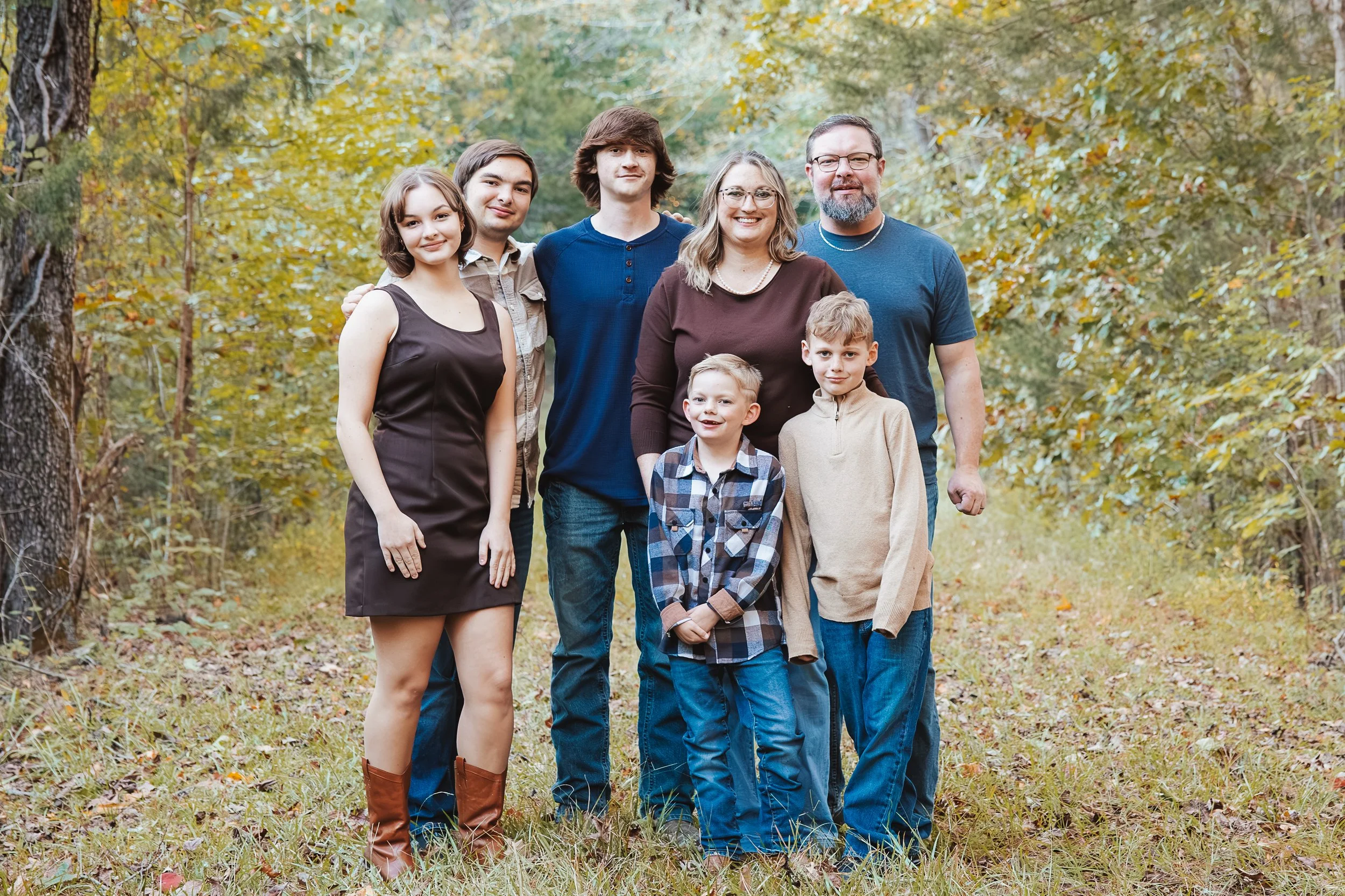 A family of seven standing outdoors in a wooded area during autumn, smiling at the camera. The family includes three adults and four children, with fall foliage in the background.