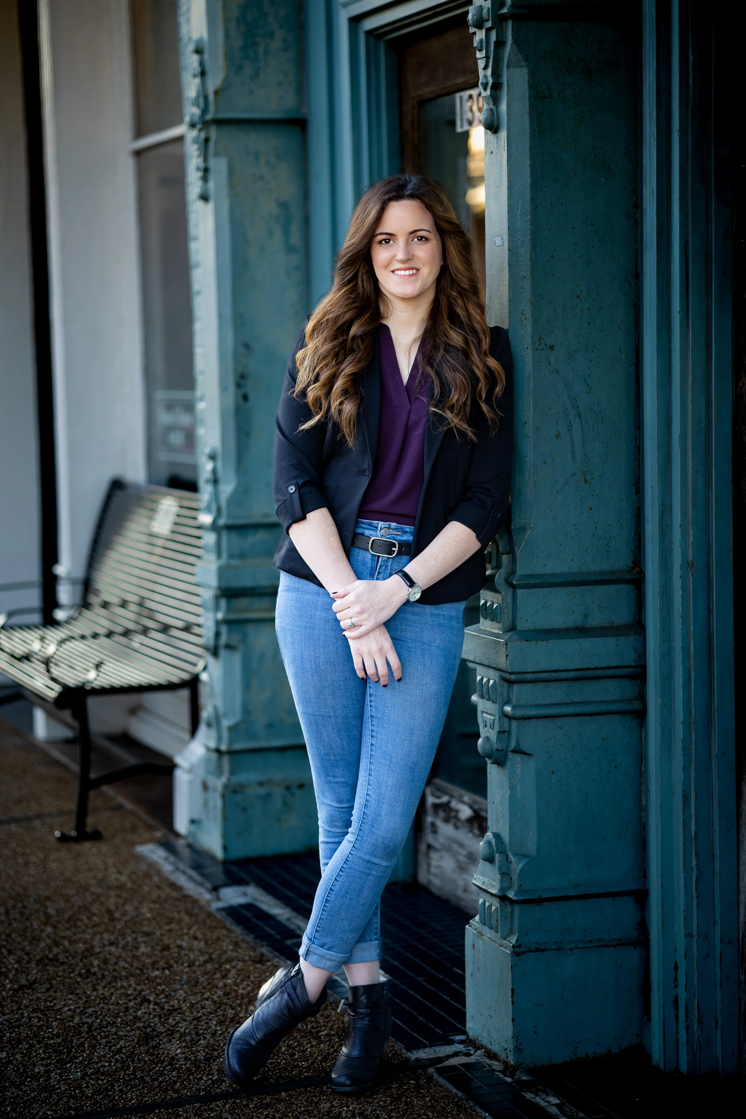 A woman with long brown hair standing outside a building with teal-colored architectural details. She is smiling, wearing a purple blouse, black jacket, light blue jeans, and black ankle boots.