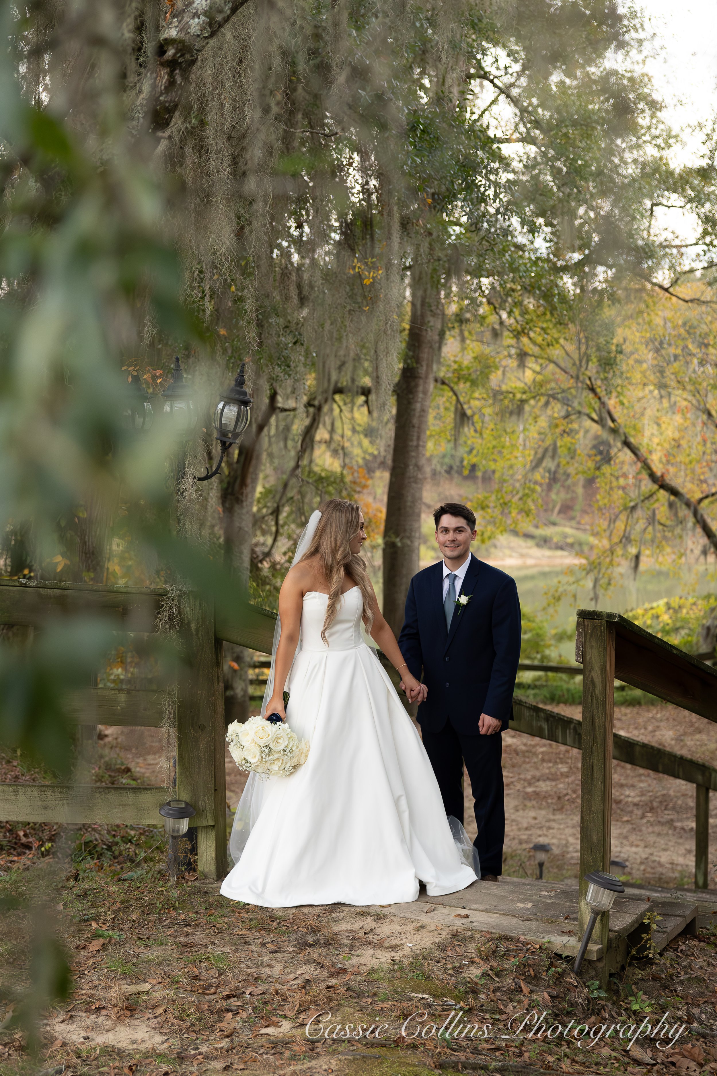 A bride and groom standing hand in hand on a small wooden bridge outdoors during a wedding photo session. The bride is wearing a white wedding gown holding a bouquet of white roses, and the groom is dressed in a dark suit with a light blue tie and a white flower boutonniere. The background features a wooded area with trees and a body of water.