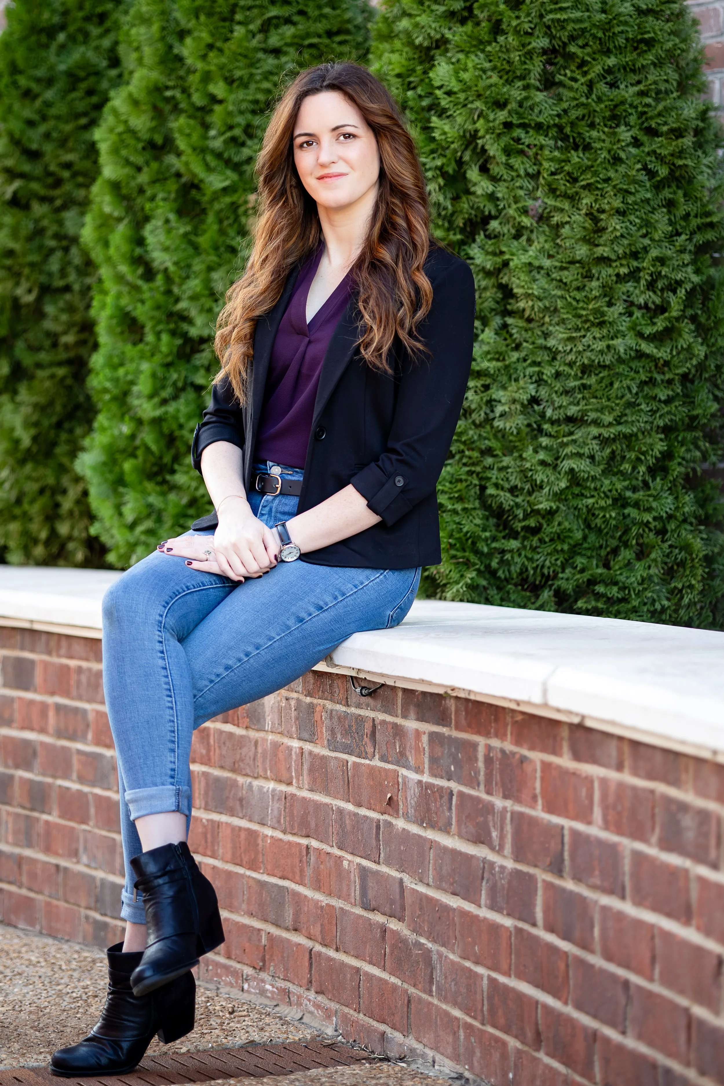 A woman with long, wavy brown hair sitting on a brick wall outdoors, wearing a black blazer, purple blouse, blue jeans, and black ankle boots, with a green leafy background.
