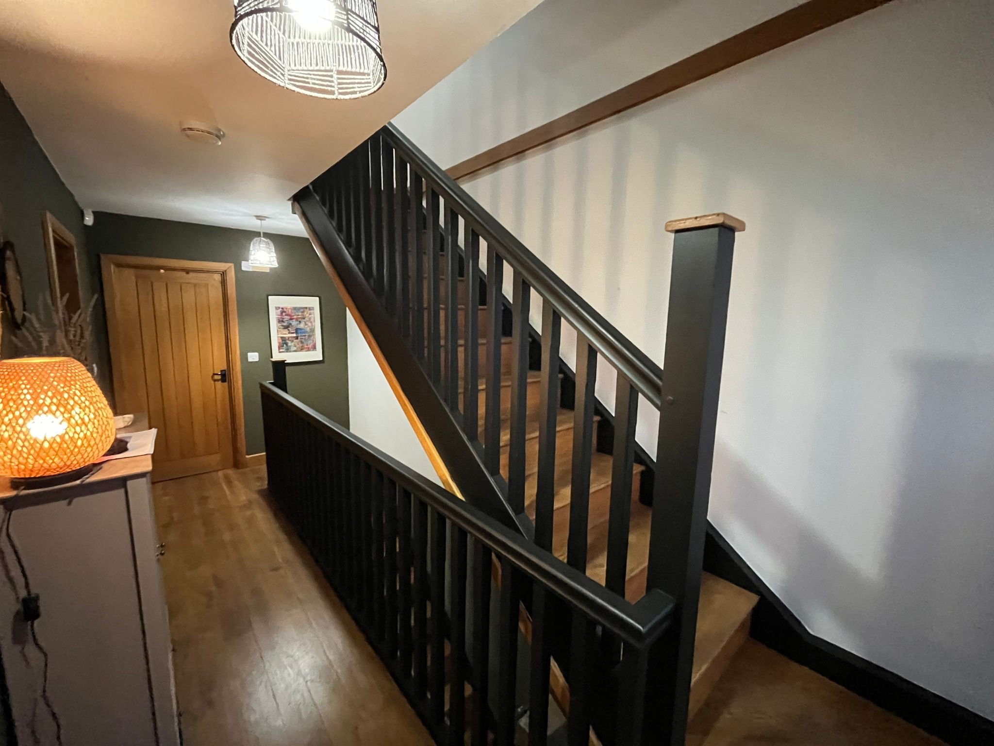 Interior view of a home staircase with black railing, wooden stairs, and wall art, in a hallway with wooden flooring and a green wall, lit by ceiling lights and a lamp.