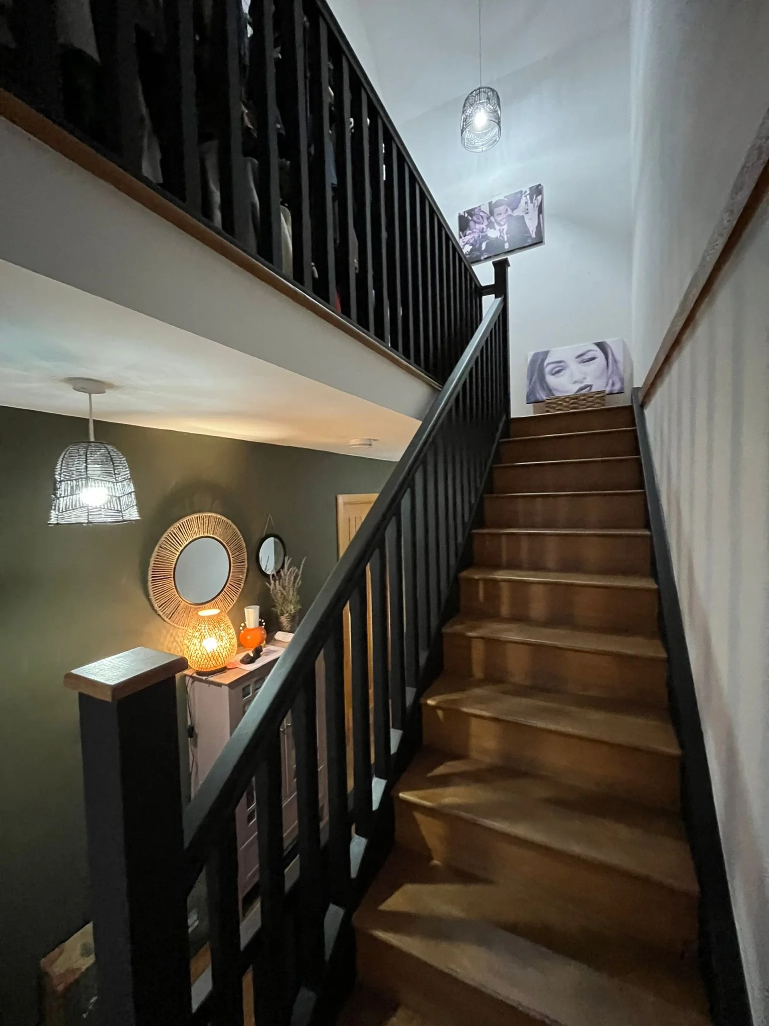 Interior view of a staircase in a home, with black railing, wooden steps, and artwork on the upper wall. A decorative lamp and a vase are visible in the lower background.
