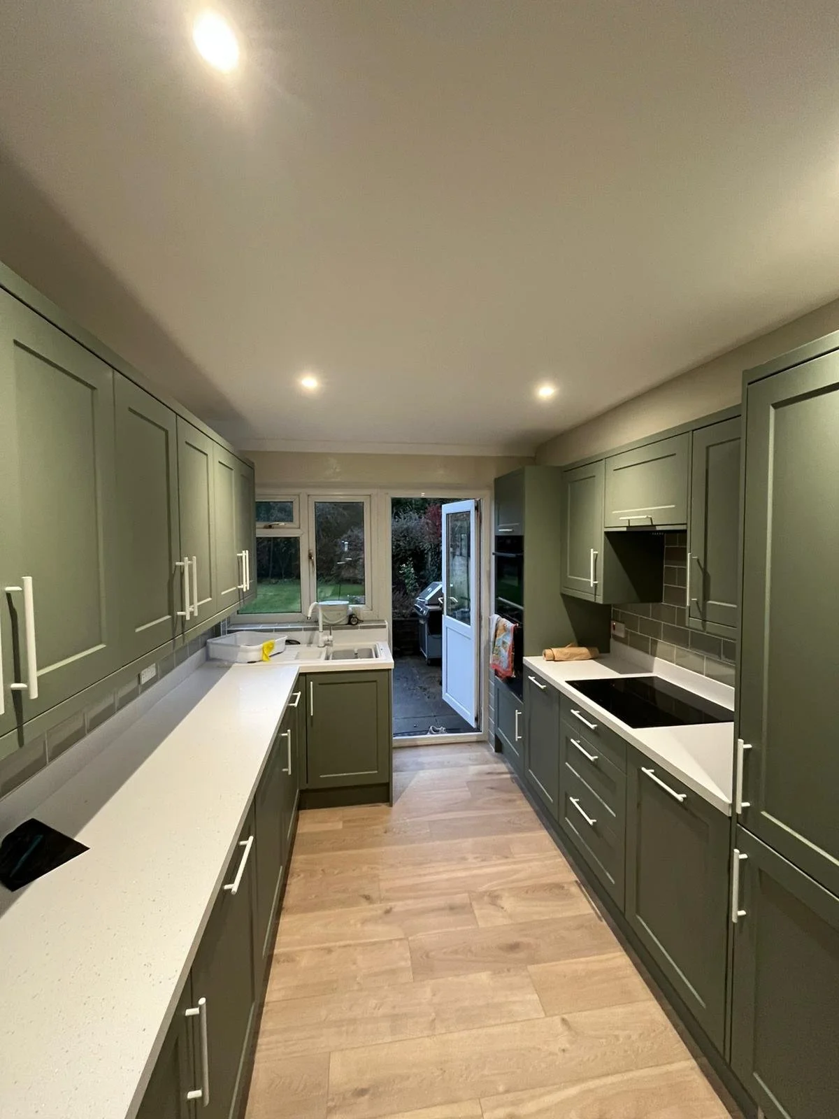 Green kitchen with white countertops, wooden floor, and open door leading outside, with a view of a garden.