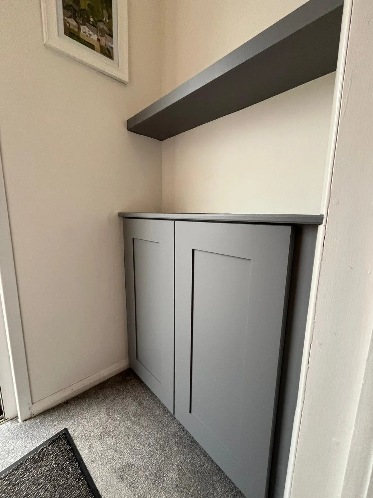 A grey cabinet with two doors, topped with a grey shelf, installed in a small corner next to a beige wall with a framed picture hanging above.