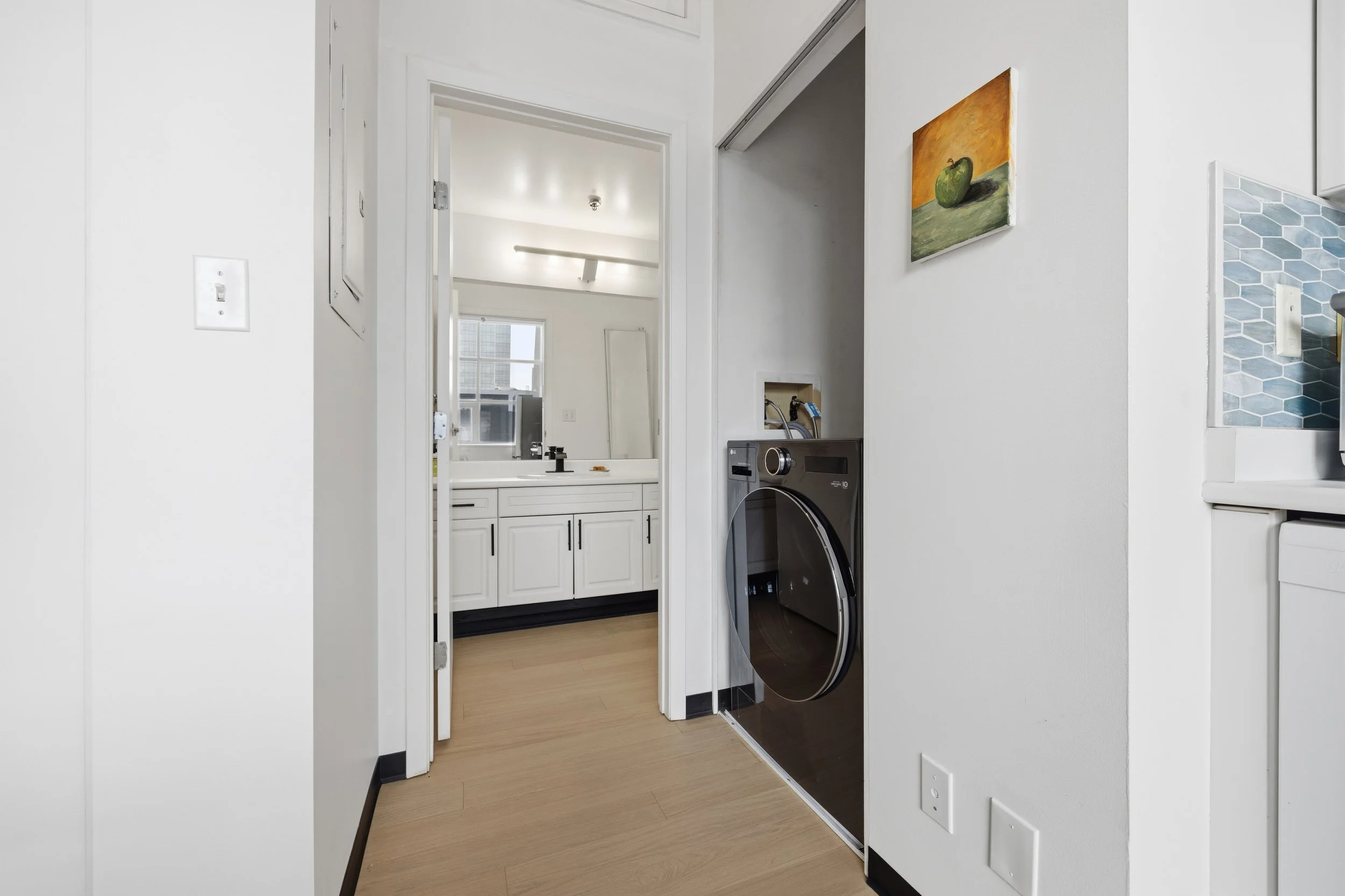 View of a laundry area with a front-loading washing machine, a white bathroom vanity with a sink, and a wall art of a green apple on the wall.