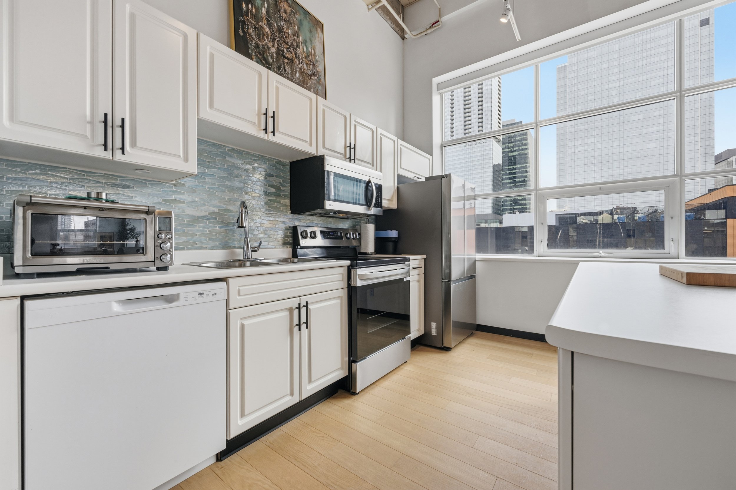 Modern kitchen with white cabinets, stainless steel appliances, a large window with a cityscape view, and a light wooden floor.
