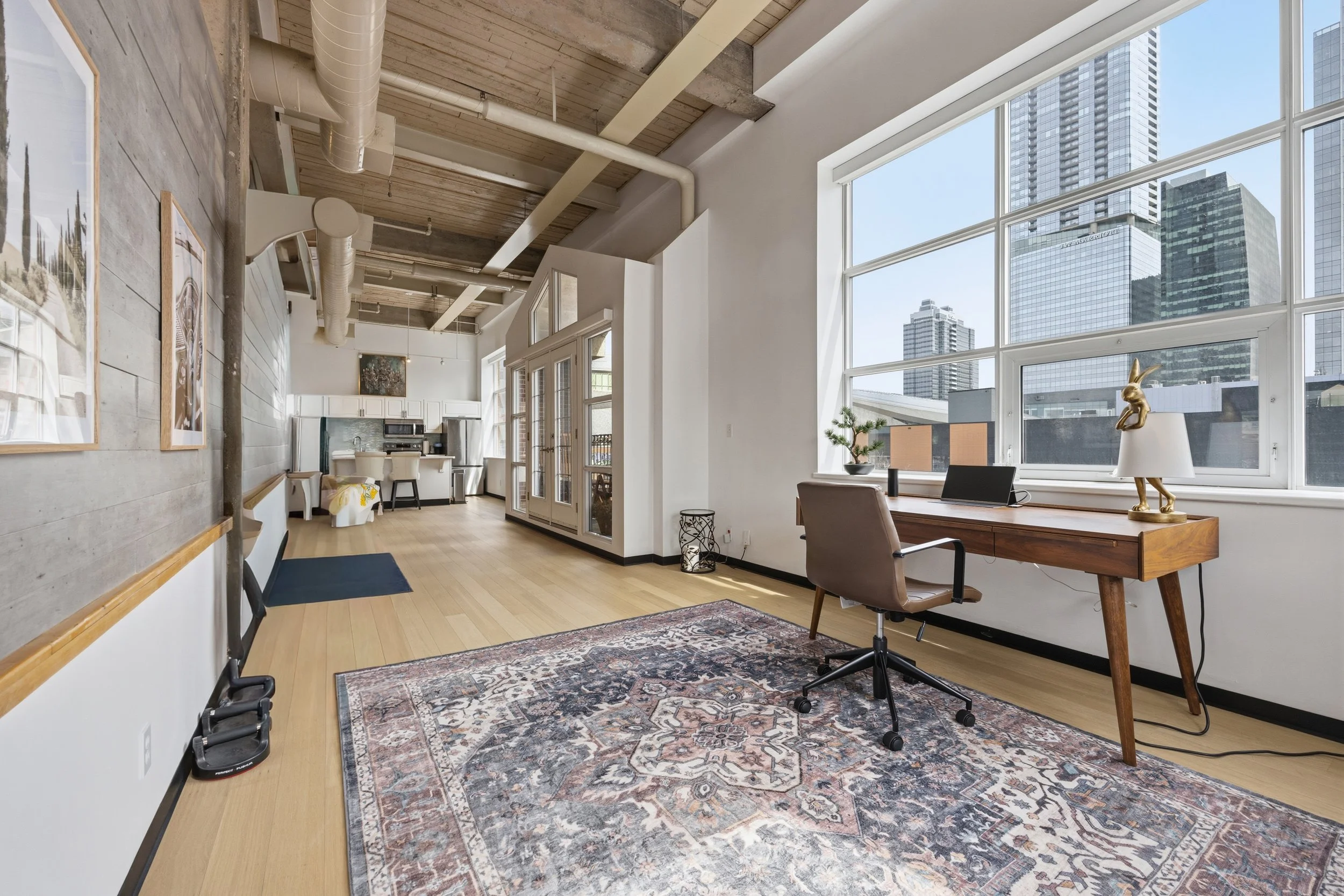 A spacious modern apartment living room with large windows showing city skyscrapers, a wooden desk with a chair and decorative items, a partial view into the kitchen and dining area, hardwood floors, and artwork on the wall.