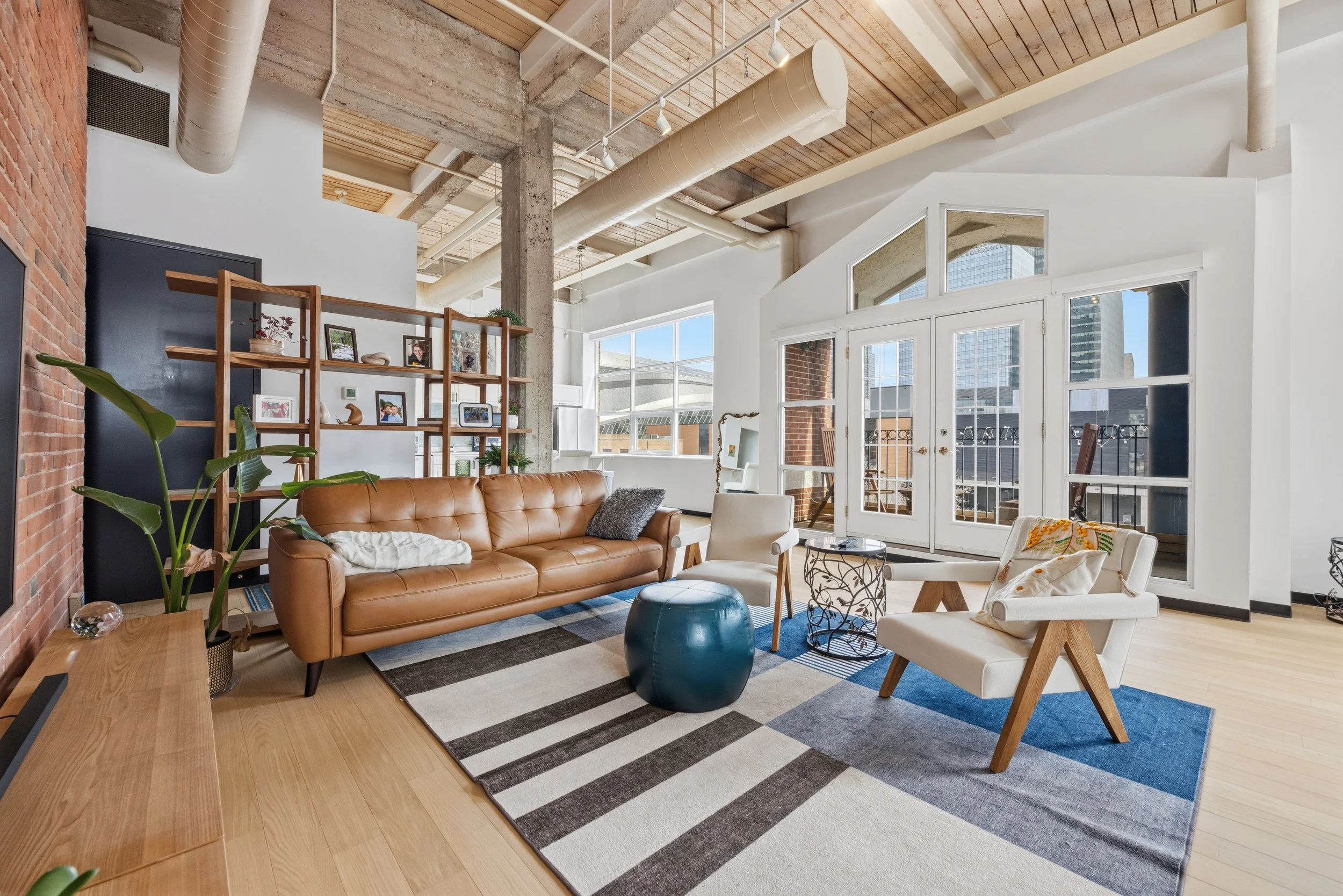 Modern living room with exposed brick wall, wooden ceiling beams, large windows, tan leather sofa, white armchairs, blue ottoman, area rug, and wooden shelving filled with framed photos and decorative items.