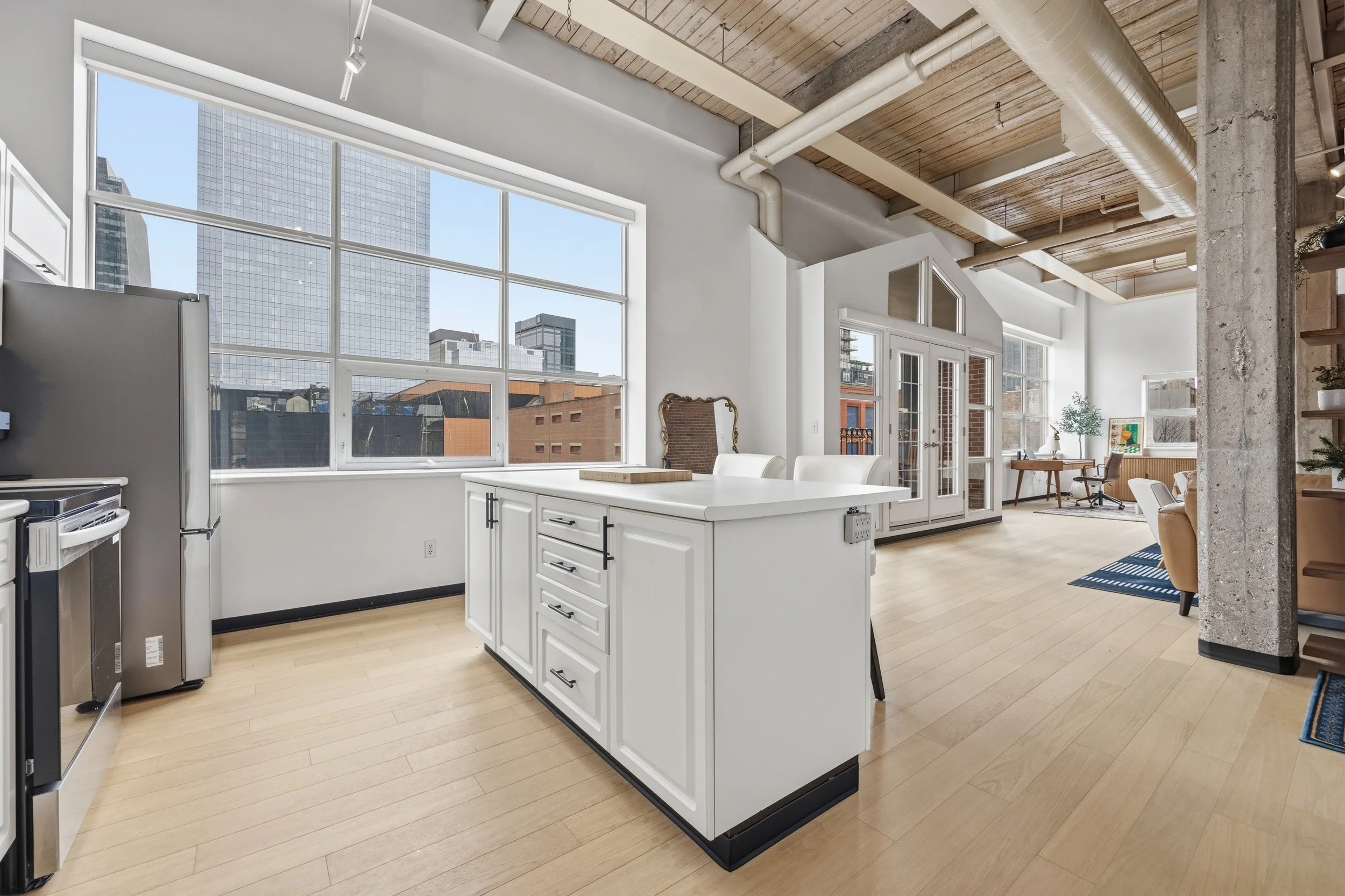Modern loft apartment interior with large windows, white kitchen island, wooden floors, and exposed ceiling beams with visible ductwork.